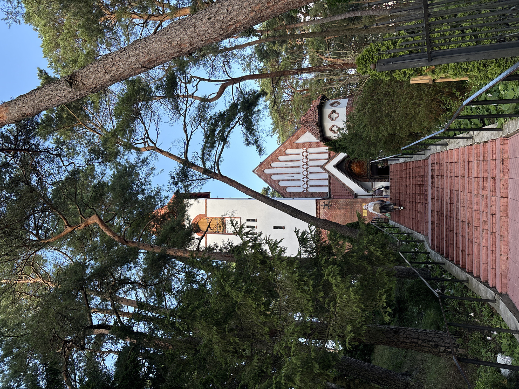 In Svetlogorsk, Russia, a person walks up a red brick staircase towards a white church with a red tiled roof, nestled amongst tall pine trees. The church features a white tower with a rounded top and a distinctive arched entrance. Two other people are visible near the entrance. The scene is set against a clear blue sky, with the lush green foliage of the surrounding trees framing the architecture.