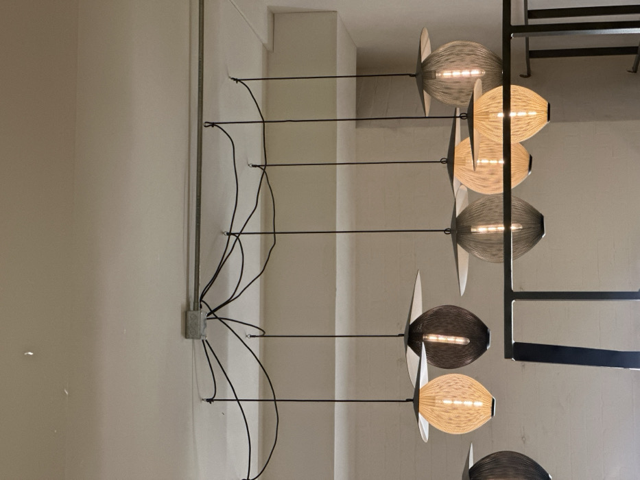 The image displays a modern interior architectural feature consisting of a series of decorative pendant lights arranged vertically on a light-colored wall. The lights are suspended from thin, dark horizontal rods or wires that extend from the wall.
On the left side of the image, a light cream-colored wall is visible. A vertical grey metal conduit runs down the wall, connecting to a square grey electrical junction box located roughly in the middle of the wall's height. From this junction box, several black electrical cords branch out, secured to the wall at various points with small metal loops or hooks. These cords run horizontally to supply power to the individual light fixtures.
There are at least seven visible light fixtures, with possibly more extending out of frame at the bottom. Each fixture consists of an elongated, somewhat oval or cocoon-shaped shade made of a finely woven or textured material, resembling string or thin wire. Inside each shade, multiple linear LED light sources are visible, emitting a warm glow. Each shade is flanked by two flat, wing-like or fin-like elements, typically lighter in color (white or very light grey) and oriented perpendicularly to the main shade.
The light shades alternate in color: one is a light beige or off-white, and the next is a darker grey or possibly dark brown/black. This alternating pattern creates a visual rhythm. The light fixtures are evenly spaced vertically, creating a continuous column of illumination.
To the far right of the light fixtures, a dark, angular metal structure, possibly part of a frame, shelf, or another architectural element, is partially visible. The background behind the lights appears to be another light-colored wall or ceiling. The lights are illuminated, suggesting they are turned on, and contribute to the ambient lighting of the space. There are no people or discernible text in the image.