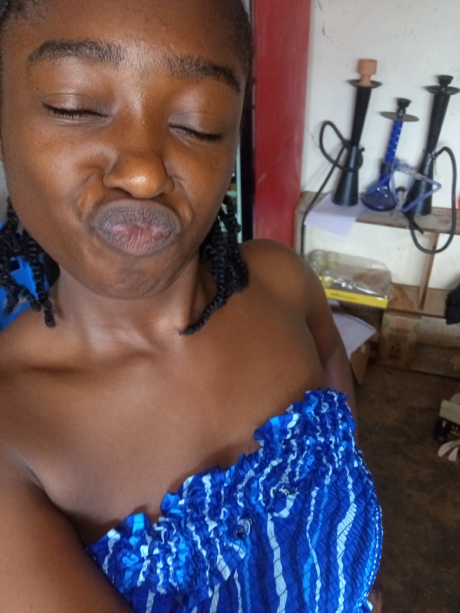 A young Black female is seen from the chest up, posing for a selfie with her eyes closed and lips puckered. She has dark skin, braided hair, and is wearing a blue and white striped strapless top. The background is cluttered with various objects, including what appear to be hookahs and some boxes. The lighting is natural and somewhat dim, suggesting an indoor setting. The overall impression is informal, possibly in a residential or casual environment.