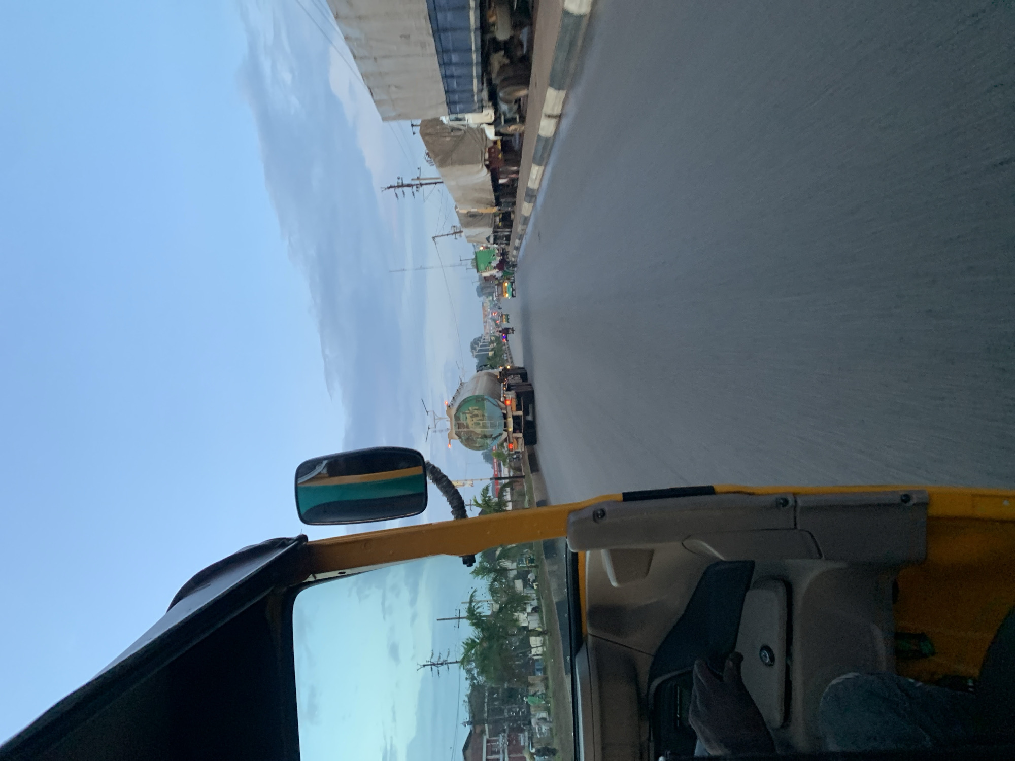The image, taken from the perspective of a passenger in a yellow auto-rickshaw in Jos, Nigeria, captures a view of a road during what appears to be late afternoon or early evening. The sky is a muted blue with scattered clouds. To the left, a portion of the auto-rickshaw's interior is visible, including a steering wheel and a side mirror. Beyond the rickshaw, palm trees and buildings line the side of the road, with several people walking and a few vehicles present.
The main focus of the image is the road itself, which is paved and appears to be a multi-lane highway. A significant number of large trucks are present, some of which are parked along the side of the road. One truck in the mid-ground is a tanker truck with a cylindrical container. Further down the road, traffic is visible, including other trucks, a few smaller vehicles, and motorcycles. The road surface shows signs of wear and tear. The overall scene suggests a busy transportation route, possibly on the outskirts or within a commercial area of Jos. The lighting indicates either the early morning or late afternoon, given the soft light and the clear sky with some cloud formations.
