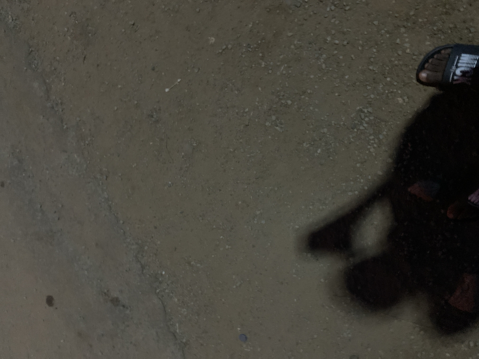 The image depicts a patch of ground in Gbeleko, Nigeria. The surface appears to be a mix of dirt and small stones. In the upper right corner of the frame, there is a person's foot wearing a sandal. The shadow of the person is also visible, suggesting a light source is present. No other people, objects, or actions can be identified within the frame.