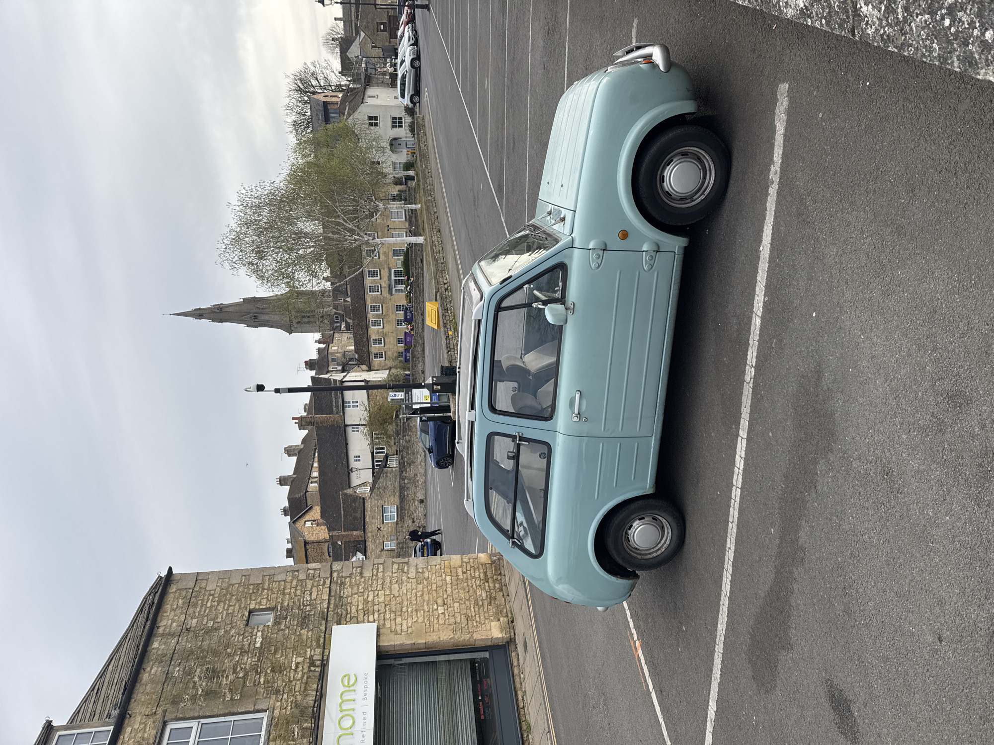 The image captures a pale blue Nissan Pao parked in a marked parking space in South Kesteven, United Kingdom. The parking lot is located in a town with a historic appearance, featuring stone buildings and a church spire visible in the background. A storefront with the sign "home" is visible to the left of the parked car. Other vehicles can be seen parked further down the row. The weather appears overcast.