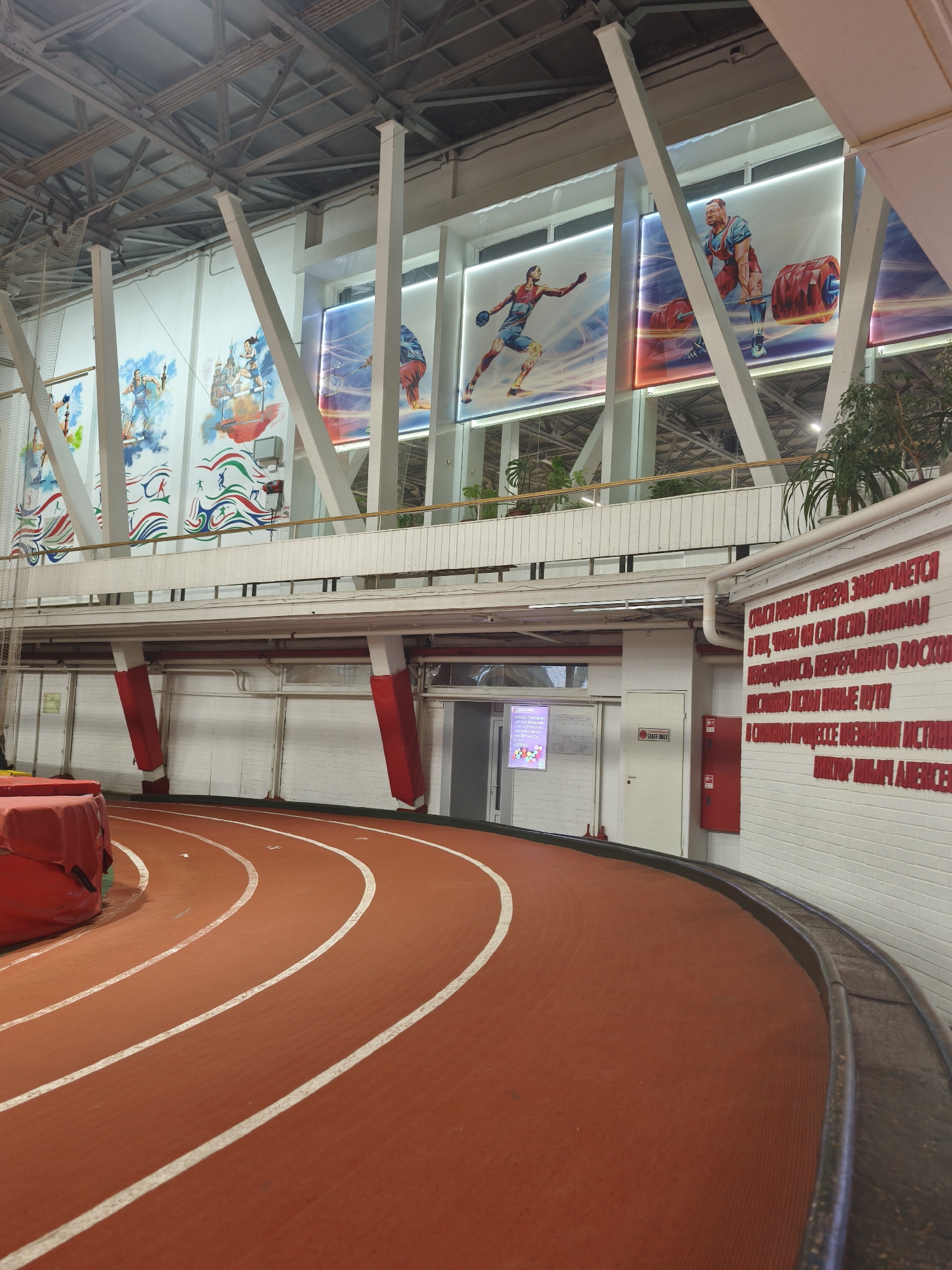 The image captures the interior of an athletic facility, specifically an indoor running track. The track itself is a reddish-brown synthetic material with white lane markers, curving towards the left. A red protective pad sits adjacent to the track.
Above the track, the walls feature large murals depicting athletes, set in illuminated frames. These murals are interspersed with white structural support beams and a white painted railing. There is a doorway marked "STAFF ONLY" on the wall. The wall is constructed of white brick.
To the right of the doorway, there is a sign with Russian text in red lettering on the wall. The upper part of the structure includes additional support beams, creating a high ceiling with visible lighting fixtures.
The scene is located in the city of Saint Petersburg, Russia.