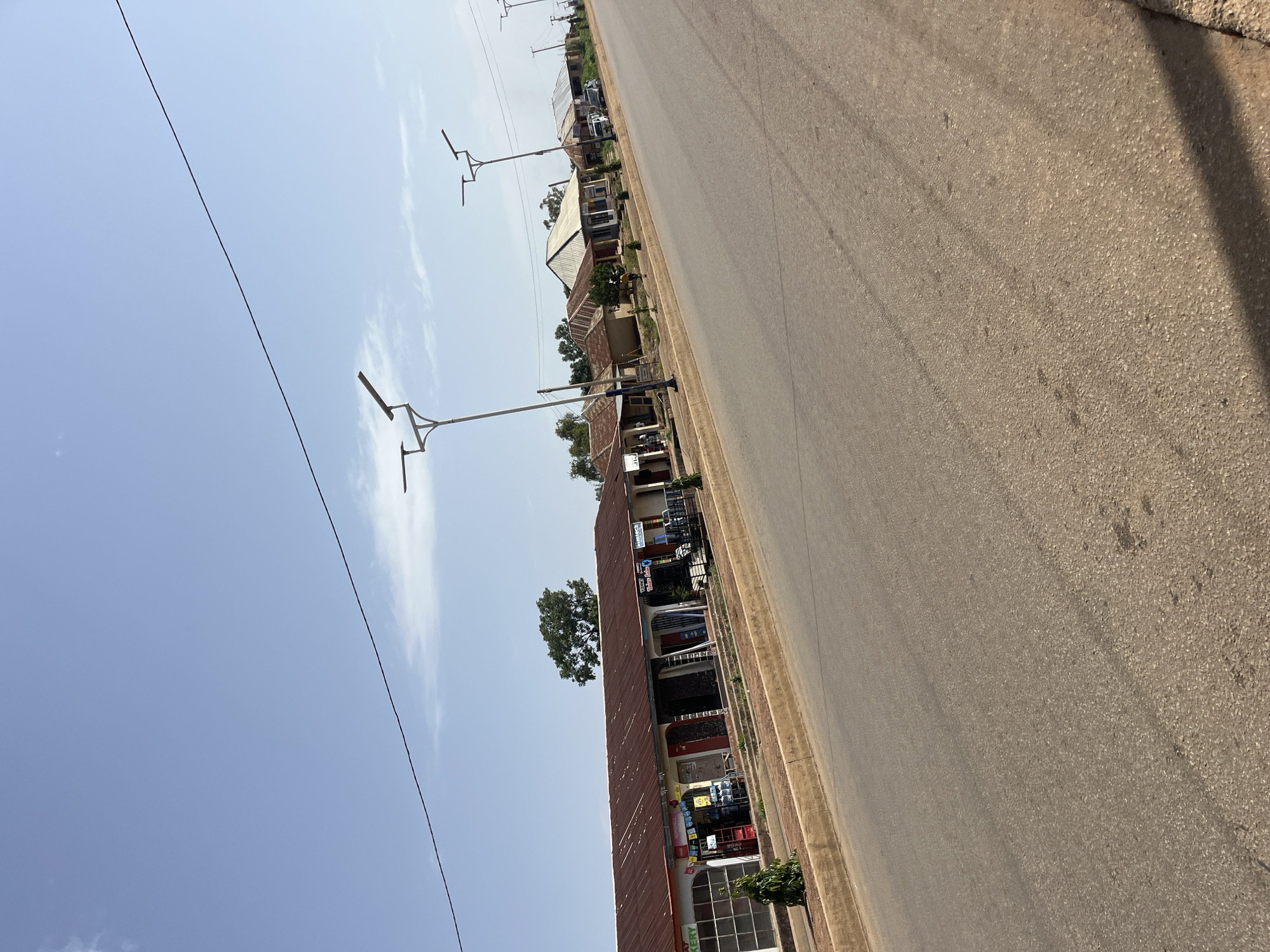A street scene in Bukuru, Nigeria, features a paved road running diagonally through the frame, lined with shops and residences on the left. The sky is a clear, pale blue with a few wispy clouds. Several modern streetlights with solar panels are visible along the road. On the left side of the street, a row of single-story buildings with corrugated metal roofs houses various businesses, including what appears to be a bakery and other storefronts with signage. Some of these shops have displays of goods outside. Further down the street, more residential-style buildings with pitched roofs are visible, some with vehicles parked nearby. Trees and shrubs are scattered throughout the scene, providing touches of greenery. The overall impression is of a typical commercial and residential street in a Nigerian town under a bright, sunny sky.