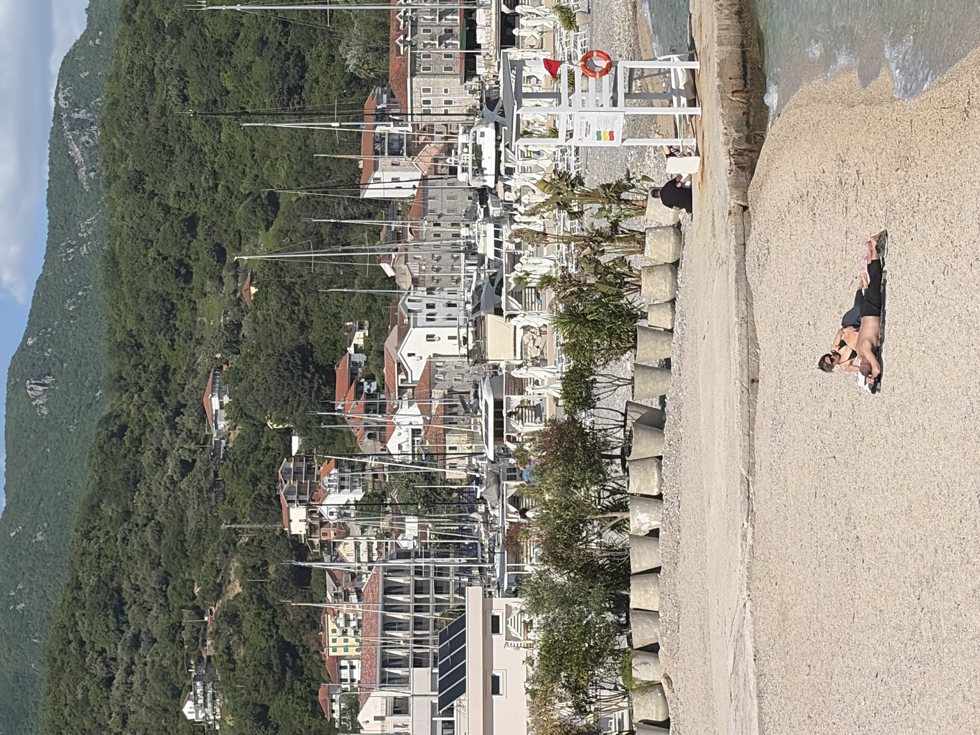 This image depicts a sunny coastal scene in Herceg Novi, Montenegro, encompassing a marina, a concrete promenade, and a lush green mountain.
In the foreground, a wide concrete path extends along the water's edge, partially transitioning into a pebbly beach where gentle waves are visible on the right. Two individuals are on this path. One person, wearing dark clothing or swimwear, is lying face down on a dark mat or towel, seemingly sunbathing or relaxing. Further left, another person, dressed in black, sits on a white chair next to a white lifeguard stand.
The lifeguard stand features a red lifebuoy and a white sign displaying red, yellow, and green flags, indicative of water safety conditions. Below these flags, there is text that is difficult to read. Another vertical white sign next to the lifebuoy also contains illegible text, likely instructions or warnings. A series of gray, cube-shaped concrete bollards line the path, interspersed with small palm trees and other green plants.
Behind the promenade, a dense marina is filled with numerous boats and yachts, their tall masts creating a prominent vertical element against the backdrop. The town's buildings, characterized by light-colored facades and terracotta roofs, stretch along the waterfront and up the lower slopes of the mountain. Some buildings have solar panels visible on their roofs.
The background is dominated by a large, heavily forested green mountain under a clear blue sky with a few faint clouds. The scene is brightly lit by direct sunlight, suggesting a clear day, likely in the morning or early afternoon. The overall impression is one of a peaceful, vibrant Mediterranean resort setting.