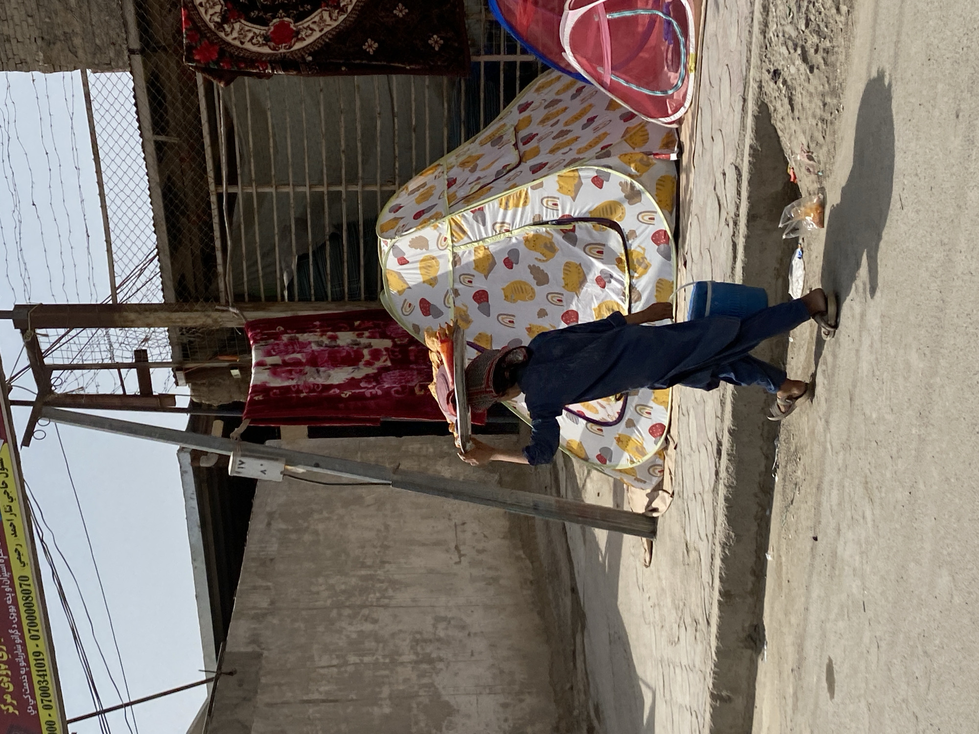 In a dusty street in the city of Kandahar, Afghanistan, a person wearing a dark blue tunic and pants walks along a sidewalk. The person is carrying a tray of food items balanced on their head and a blue bucket in their left hand. Behind them, a tent with a white base adorned with a pattern of yellow cats and rainbows is visible, alongside a smaller pink tent. Several blankets are draped over a metal structure, one with a floral pattern and another with a red and black design. The environment appears to be an outdoor market or residential area, with concrete walls and a paved street. The sky is a clear blue.