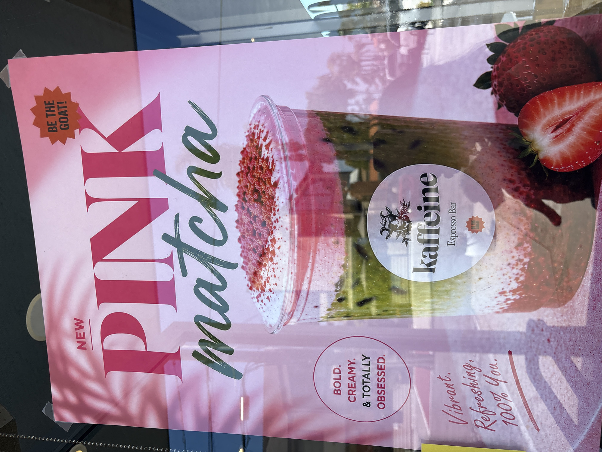 The image shows a promotional poster for "Pink Matcha" from a coffee shop named "Kaffeine Expresso Bar". The poster features a photograph of the Pink Matcha drink topped with red sprinkles in a clear to-go cup. To the right are strawberries. The text on the poster reads: "NEW," "PINK matcha," "BOLD. CREAMY. & TOTALLY OBSESSED.", and "Vibrant. Refreshing, 100% You.". The "Kaffeine Expresso Bar" logo is present as well. There is a promotional sticker that reads, "BE THE GOAT!". The poster appears to be displayed in the window of the coffee shop. The window is a bit reflective, so there is an image of trees and the reflection of another building. The reflection of the trees hints at the location of Redondo Beach, United States of America.
