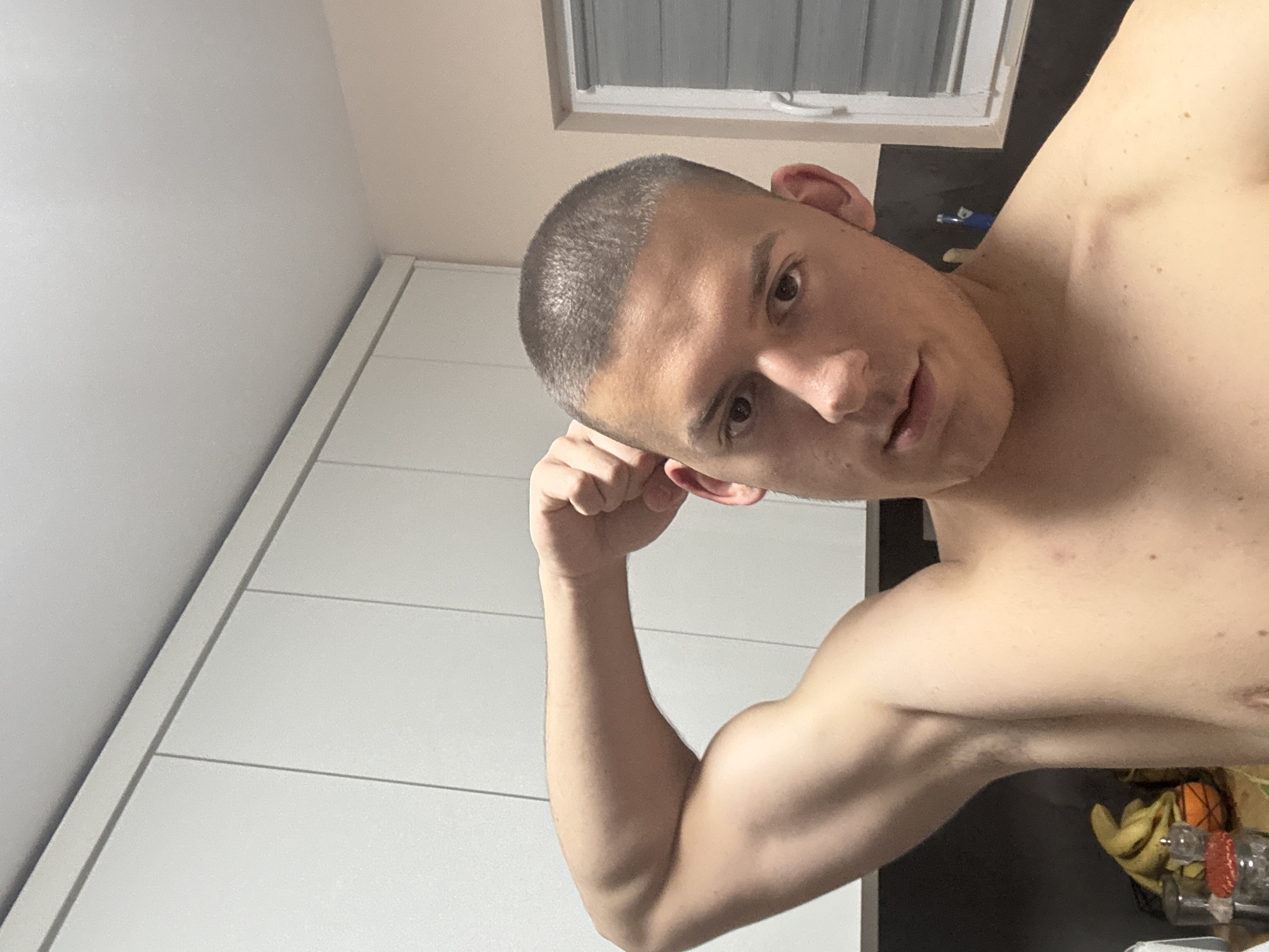 A shirtless man with a buzz cut is posing for a selfie. His left arm is flexed, showing off his bicep. He is in a room with white cabinets and a window. The location cannot be determined as Burgas, Bulgaria.