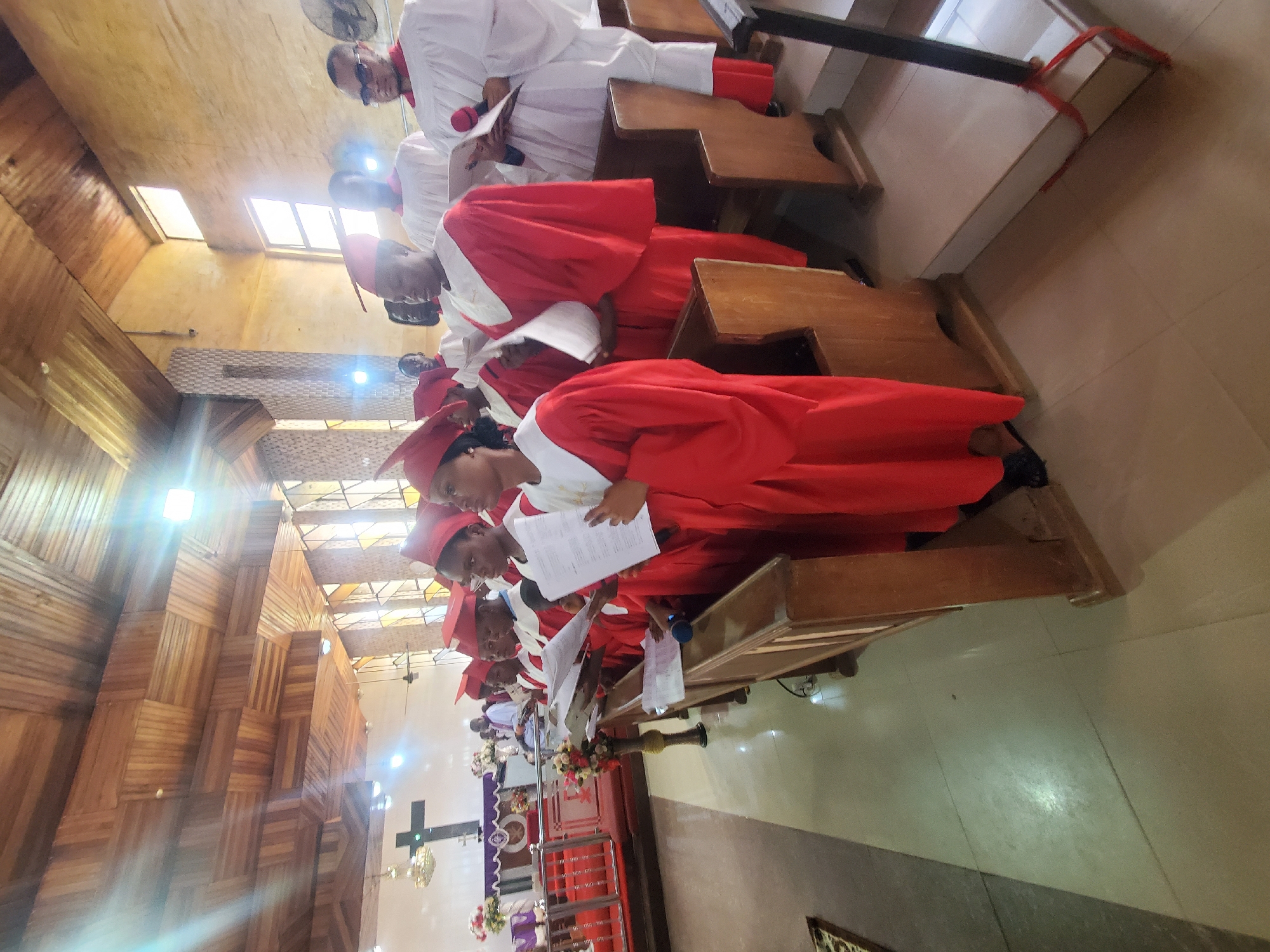 The image displays an indoor scene within what appears to be a church or a formal hall. A group of people, primarily young individuals, are seated in wooden pews. The most prominent subjects are several young people dressed in matching bright red academic-style gowns and mortarboard hats, often with white stoles or collars. Some individuals, particularly adults further back, are wearing white robes.
The activity taking place seems to be a formal gathering, possibly a church service, a choir performance, or a graduation ceremony. Many of the participants are holding white papers or booklets, which they appear to be reading or following along with, suggesting the singing of hymns, recital of prayers, or participation in a program.
Notable details of the setting include traditional wooden church pews arranged in rows. In the background, there is an altar or stage area with a large black cross and floral arrangements. Parts of the walls and ceiling feature intricate wooden paneling. Natural light streams in from windows in the upper section of the room, creating bright highlights and lens flares, suggesting it is daytime. Artificial overhead lighting is also visible. The floor is tiled. The expressions of the individuals are generally attentive and focused. No specific text on the papers held by the participants is legible.