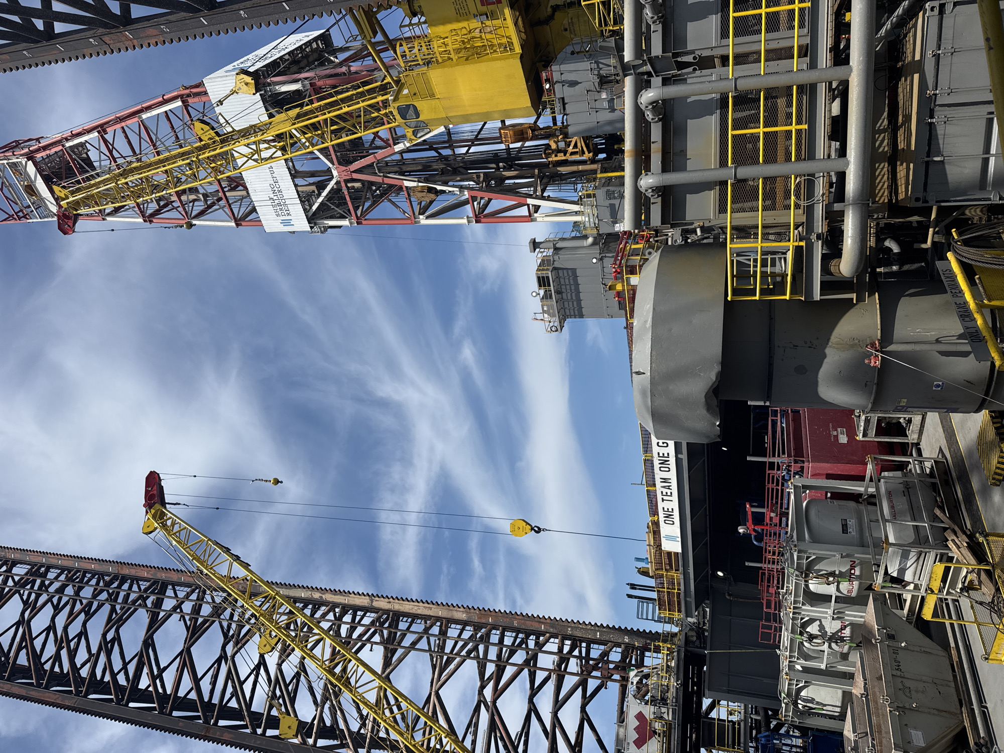 This image shows an industrial scene with a large oil rig in the foreground and a clear blue sky with scattered clouds above. The oil rig features a tall, red and white structure with a yellow crane extending from it. The rig is comprised of multiple levels of metal platforms and pipes, with various equipment visible, including large grey tanks and yellow safety railings. A prominent sign on one of the platforms reads "ONE TEAM ONE G". The overall impression is of a busy industrial operation on a sunny day. There are no people visible in the image. The location is not specified as being in the city of Unknown, Italy.