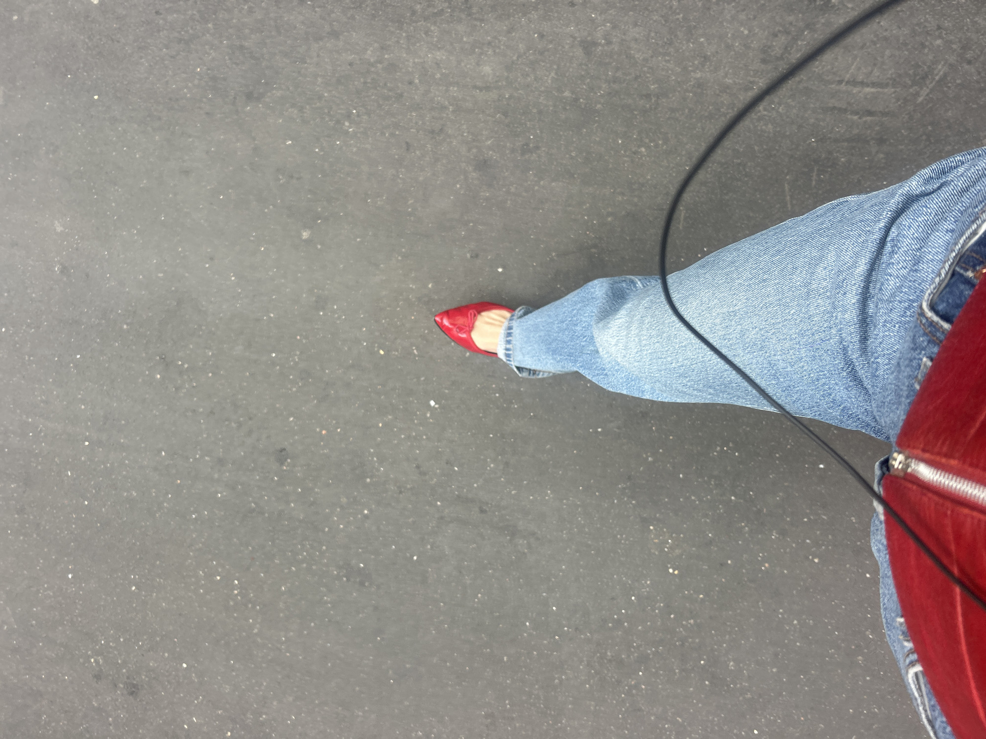 An overhead shot captures a person's lower body, specifically their right leg and foot, against a dark grey, textured surface that appears to be pavement or asphalt. The person is wearing light blue denim jeans with a rolled cuff, and a bright red, pointed-toe flat shoe or low heel is visible on their foot. A partial view of a red object, possibly a bag or purse with a silver zipper, is seen on the far right, resting against the person's leg. A thin black cord or cable stretches diagonally from the top right of the frame towards the lower center. The dark grey ground is flecked with lighter specks, suggesting wear or small debris. The lighting is even, indicating daytime, but no specific weather conditions or time of day can be determined. There is no visible text in the image.