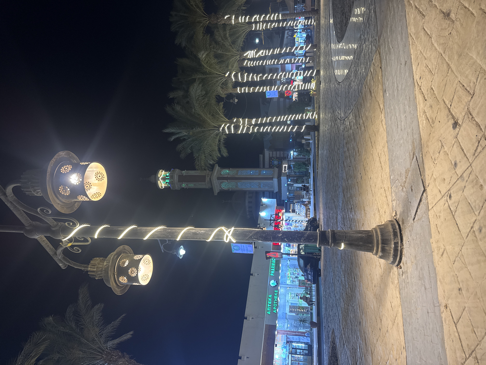 A nighttime scene in an unknown city features a street view with illuminated storefronts and decorative lighting. A prominent street lamp with ornate, perforated lanterns and string lights is positioned in the foreground. Palm trees, also adorned with string lights, line the street in the background. A building with a minaret can be seen further down the street. One of the storefronts has the word "APTEKA" displayed, indicating a pharmacy. The street is paved with light-colored brick or stone.