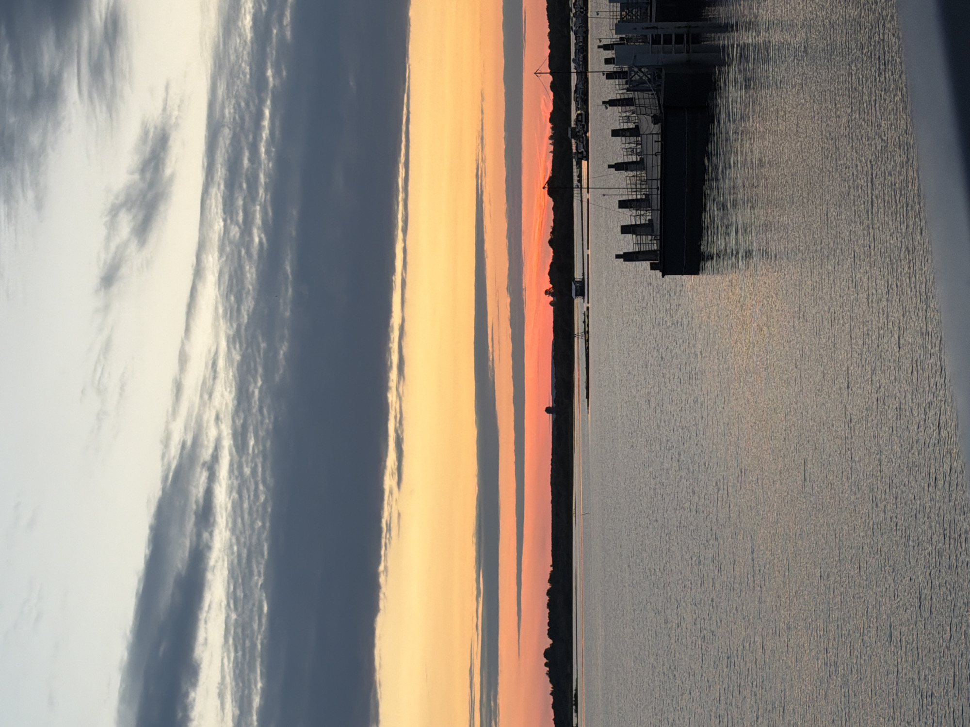 This image depicts a serene sunset over a body of water, likely a river or a lake. The sky is a vibrant gradient of colors, transitioning from a pale yellow and orange near the horizon to darker blues and grays at the top. Streaks of clouds, some dark and ominous, others wispy and light, are scattered across the sky.
In the distance, a dark, silhouetted treeline forms the horizon. On this landmass, a few structures are faintly visible, including what appears to be a water tower or a domed building. The water itself is calm, reflecting the colors of the sky and showing subtle ripples.
In the foreground, a portion of a dock or pier is visible on the right side of the image. Several dark, vertical posts rise from a dark, flat structure, suggesting a mooring area for boats. A few boats are also visible docked in the middle ground on the right.
This scene is identified as the Solikamsk Reservoir in Solikamsk, Russia. The overall atmosphere is one of tranquility and natural beauty, capturing the fleeting moments of a sunset. There are no people or discernible human activity depicted in the image, focusing solely on the landscape and the natural light.