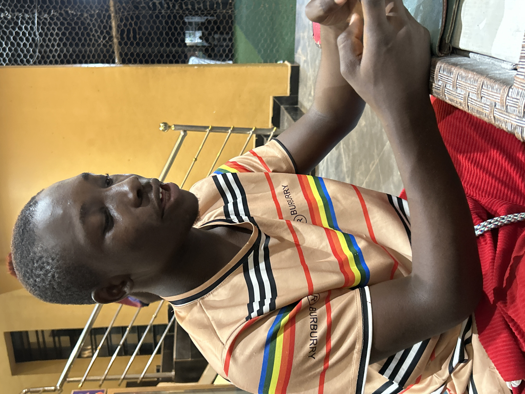 A young Black male with short, dark hair is seated at a table in what appears to be an indoor recreational facility. He is wearing a tan-colored t-shirt with horizontal stripes in various colors, including red, yellow, blue, and green, as well as black and white stripes. The word "BURBERRY" is visible on the t-shirt in two locations. His hands are resting on the table in front of him. The background features a yellow wall, a metal railing with a golden ball finial, stairs, and a netted enclosure. The lighting is warm, suggesting artificial illumination. The scene is likely within Ihiagwa, Nigeria, given the context of the request.