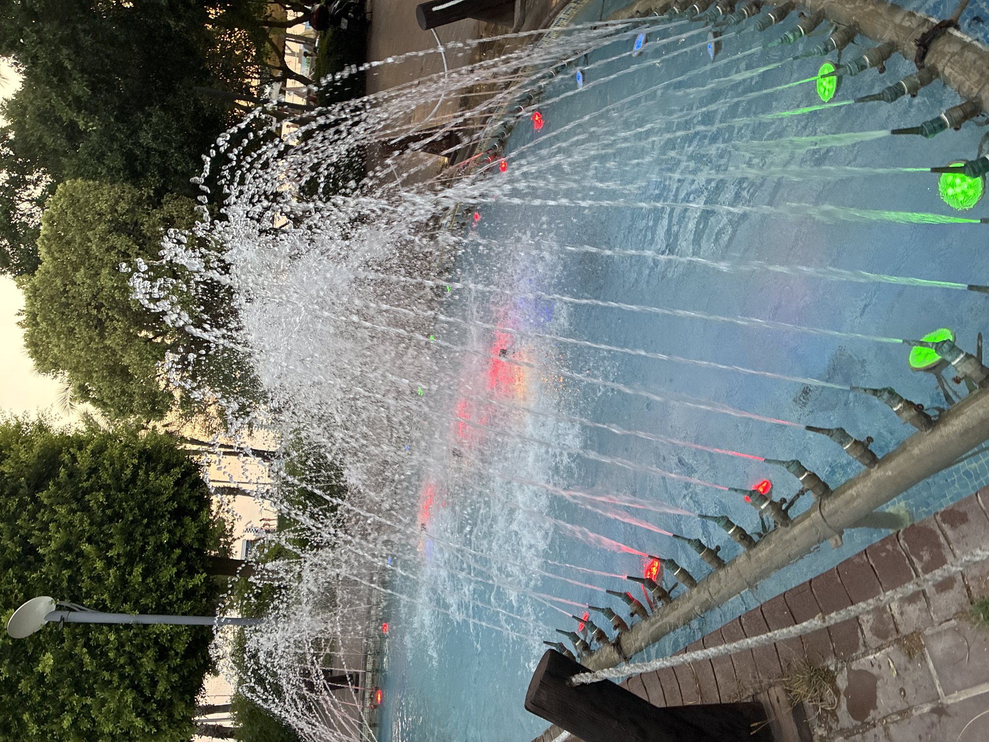 The image depicts an outdoor fountain with multiple jets of water spraying upwards and outwards, creating a dynamic visual effect. The water is illuminated from below by colored lights, with prominent red and green hues visible within the pool. The fountain is situated in a paved area, possibly a public park or promenade, with trees and vegetation forming a natural backdrop. In the background, obscured by the fountain's spray, are glimpses of what appear to be people and possibly a motorcycle, suggesting a public space in use. The lighting and the style of the fountain suggest it is either dusk or a dimly lit evening. Based on the presence of palm trees and the style of architecture and landscaping, this scene is consistent with locations in Alanya, Turkey.