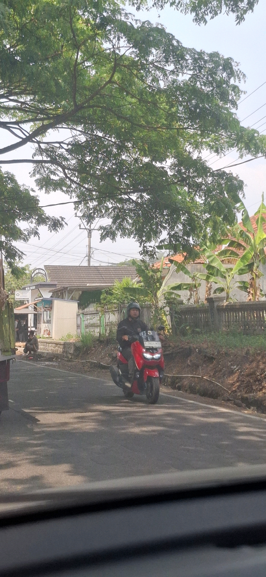 A man wearing a black jacket and helmet rides a red motorcycle on a paved road in Susukan, Indonesia. The road is flanked by a dirt embankment on the right and a fence separating houses on the left. Banana trees and other foliage are visible behind the fence. In the background, there is a building with a tiled roof and a utility pole with electrical wires. To the left of the road, a person is crouched down near a vehicle, possibly working or resting. The scene is illuminated by daylight, casting shadows from trees onto the road.