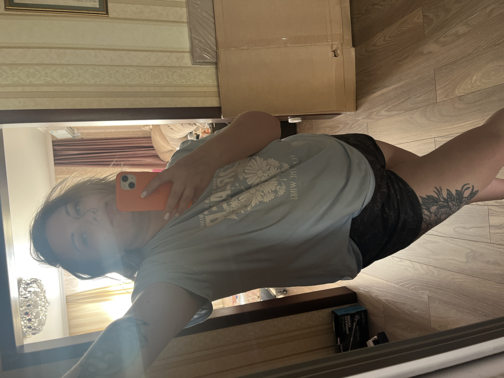 A young woman takes a selfie in a mirror. She holds an orange smartphone. The woman wears a light blue t-shirt and dark shorts. A tattoo is visible on her thigh. The mirror reflects an interior with a wooden floor, wallpapered wall, and a cardboard box. A chandelier is reflected in the mirror, as well as other interior elements. It is impossible to determine if the location is the city of Usman, Russia from the image.