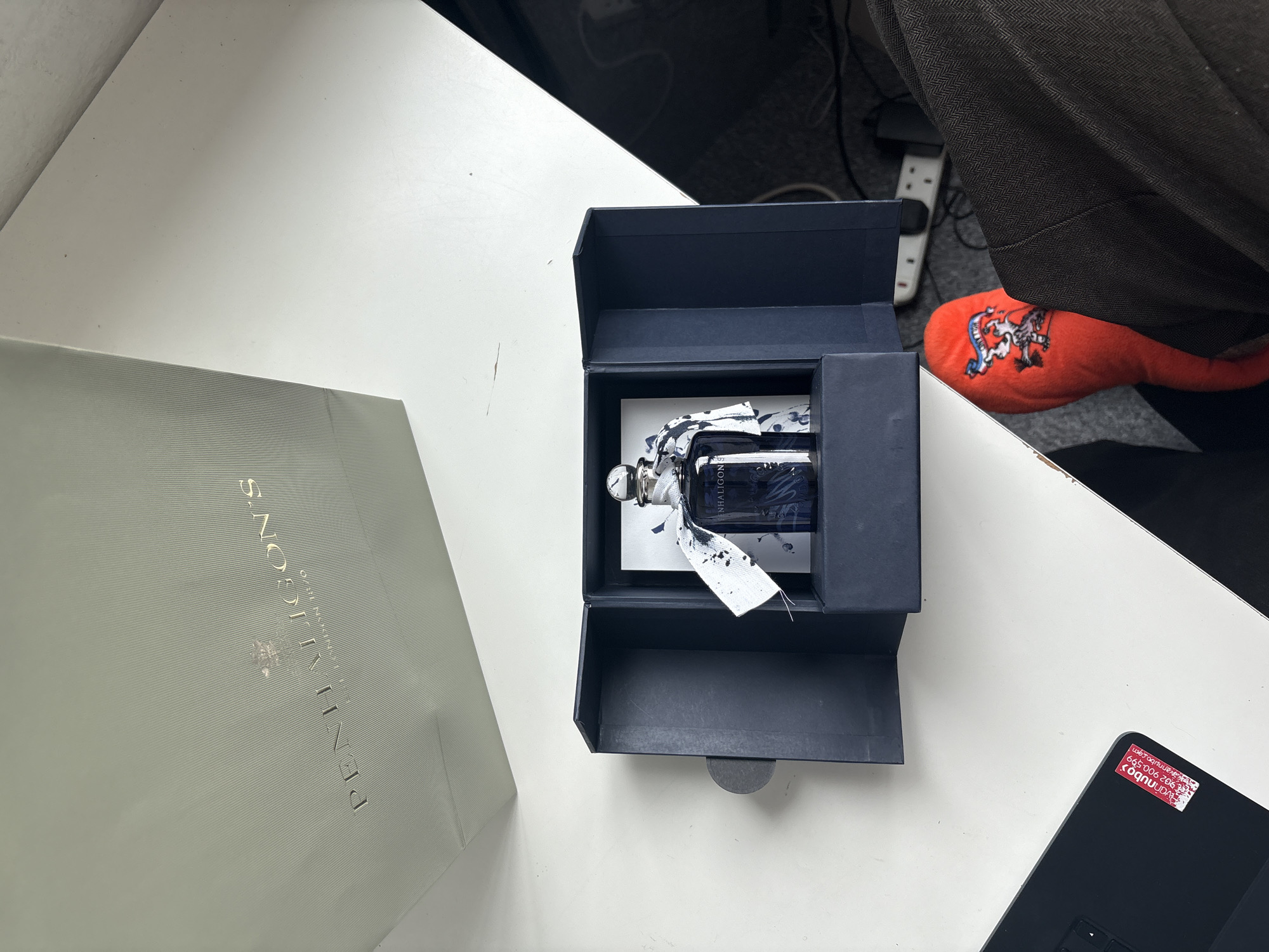 The image displays an indoor scene on a white table surface, likely in London, United Kingdom. A light gray shopping bag from "PENHALIGON'S" with a gold embossed logo and "EST. 1870" is positioned on the left. Adjacent to it, a dark blue or black presentation box lies open, revealing a dark blue perfume bottle nestled within. The bottle features a silver cap and a white ribbon around its neck, stained with dark blue or black splatters. The brand name "PENHALIGON'S" is partially visible on the bottle.
In the upper right background, part of a person's leg is visible, clad in brown trousers with a herringbone pattern. An orange plush item, resembling a slipper or soft toy, peeks from beneath the trousers, adorned with an embroidered white and blue crest depicting a lion and another animal. A white power strip with multiple sockets and a black plug is seen behind the leg.
On the bottom right, a corner of a black electronic device, possibly a laptop or tablet, shows a red and white sticker. The text on the sticker, when read upright, displays "evanunbo", a phone number "902 900 599", and "www.evanunbo.com". The overall scene suggests the unboxing or display of a luxury fragrance. The time of day is not discernible, but the area is well-lit.