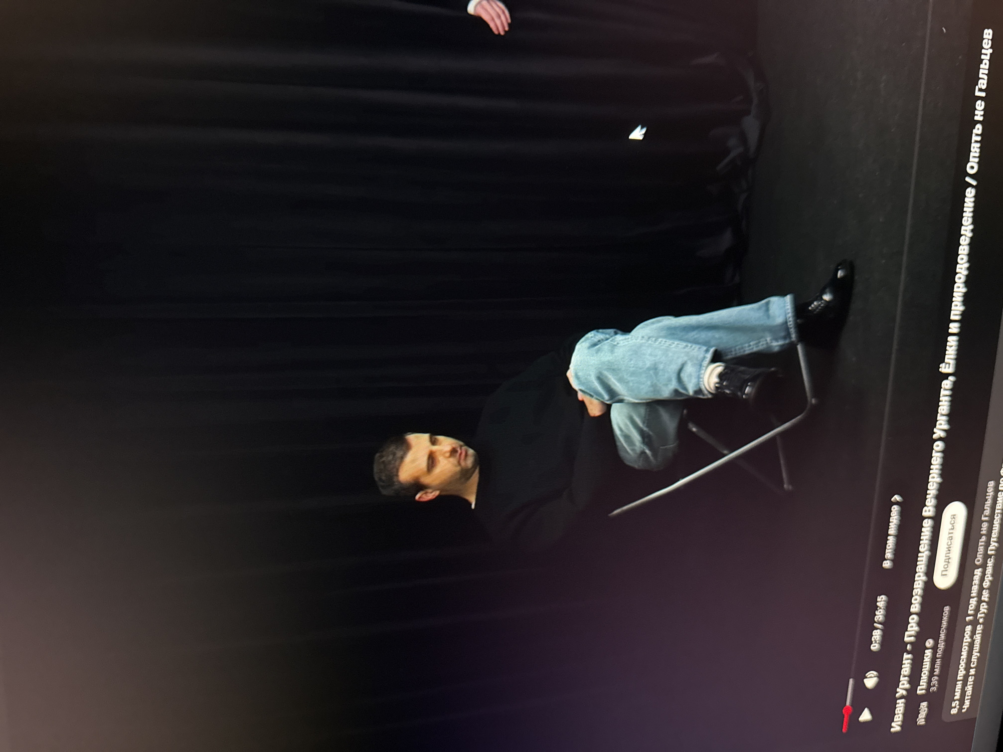 The image is a rotated close-up of a computer screen showing a paused YouTube video. The video is titled "Ivan Urgant - About the return of Evening Urgant, Christmas trees and natural history / Again, not Galtsev." A man is seated on a chair in the video. He is wearing jeans and a black shirt. The background behind him is black. The video's progress bar shows it is 38 seconds into a 36-minute 45-second video. The video has 8.5 million views and 3.39 million subscribers. A "Subscribe" button is visible. The YouTube user "Plushki" is mentioned. The location is Unknown, Unknown.
