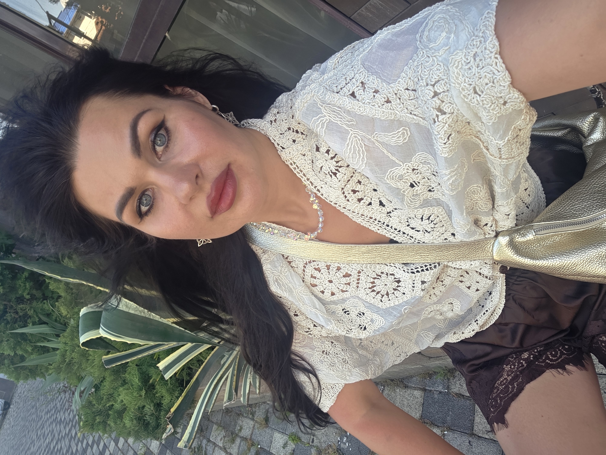 The image shows a woman with long dark hair and blue eyes, wearing a cream-colored, crocheted blouse and brown shorts with lace trim. She has a gold purse slung over her shoulder. She is outdoors, with a cobblestone path and greenery in the background. The scene appears to be a street or garden area within a city. The lighting suggests it is daytime.