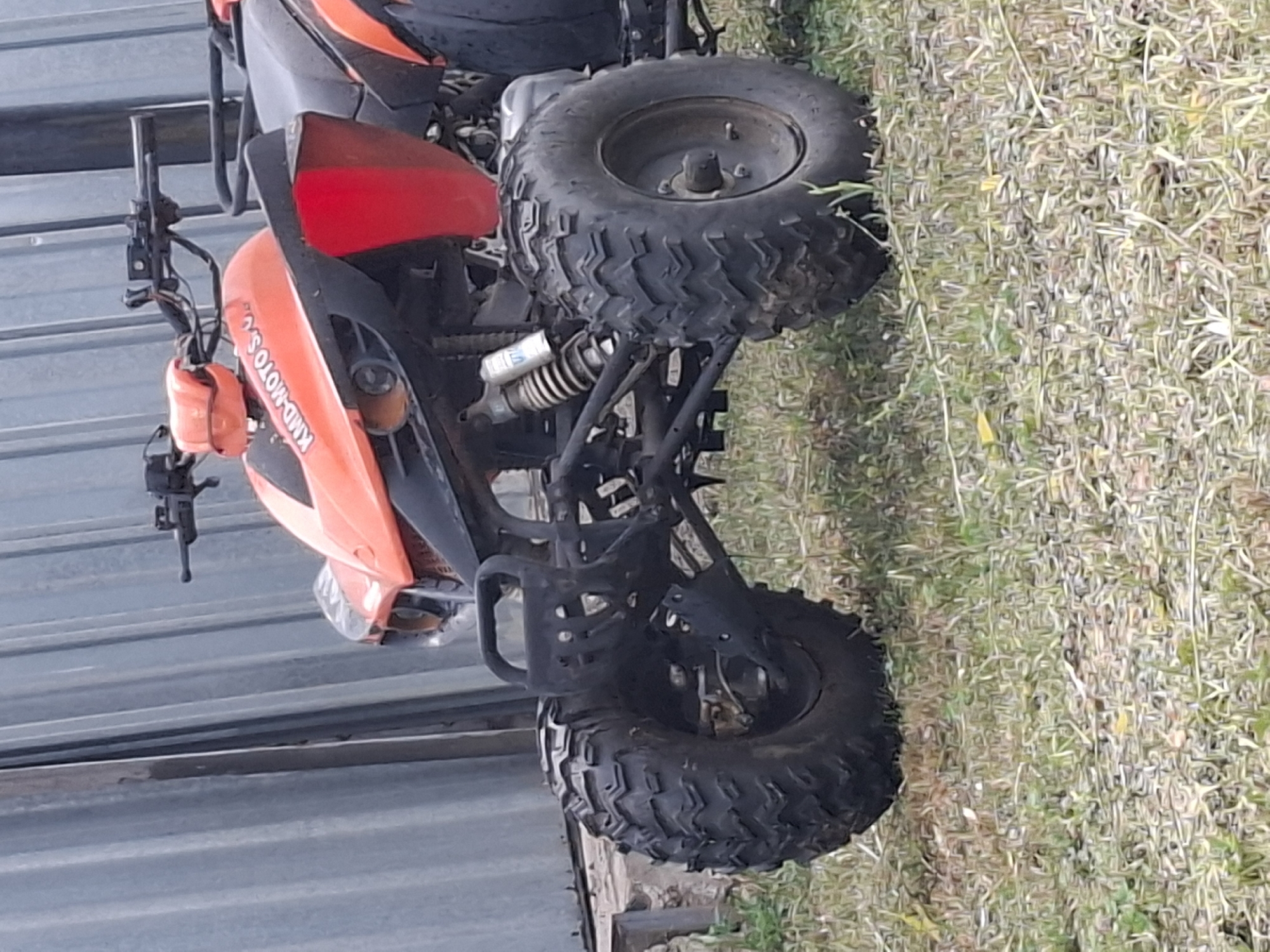 A red and black quad bike is parked on a grassy area in front of a corrugated metal wall. The quad bike has large, knobby tires and an orange front fairing with the words "KMD-MOTO S.U." visible. The handlebars, suspension, and part of the seat are also visible. The background consists of the metal wall and some dry grass. The image appears to be taken outdoors in daylight. There are no people in the image.