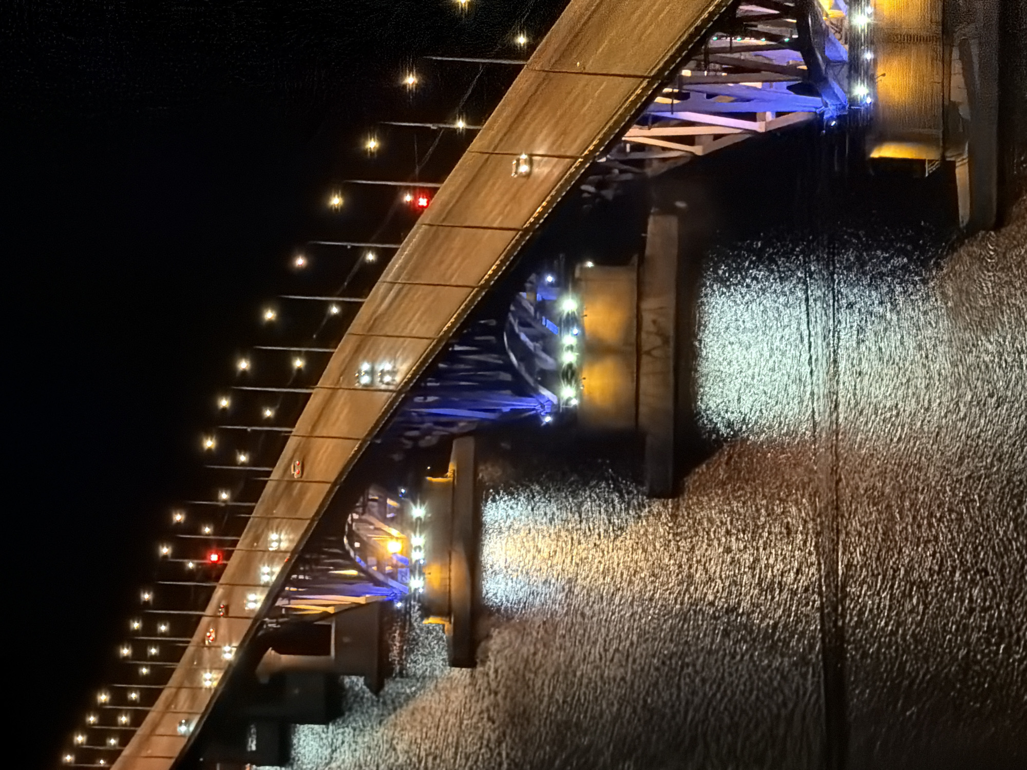 The image shows a nighttime view of the Saratov Bridge, a suspension bridge spanning the Volga River in Saratov, Russia. The bridge is illuminated by numerous streetlights, casting a warm glow on the roadway and its reflection on the dark water below. Several cars are visible on the bridge, their headlights and taillights creating streaks of light. The bridge's supporting structure is adorned with blue and yellow lights, adding to the vibrant nighttime ambiance. The water surface is textured with ripples, reflecting the lights from the bridge and creating a dynamic and visually appealing scene. The dark sky above is devoid of stars, indicating the presence of light pollution from the city. The scene is captured from an elevated perspective, providing a wide view of the bridge and its surroundings.