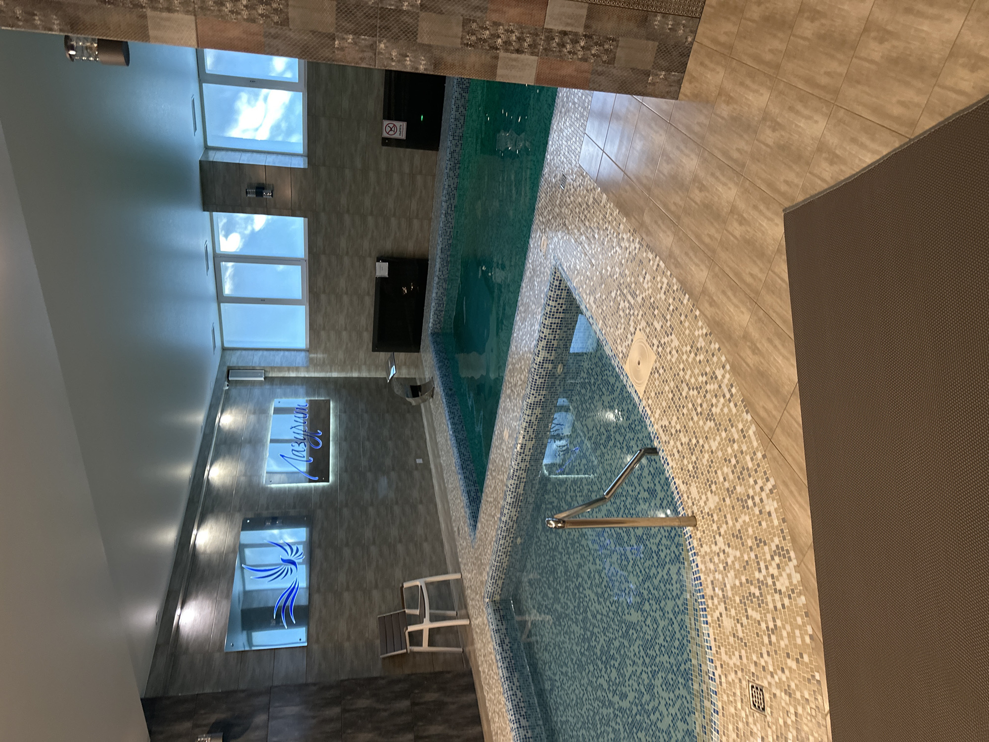 The image shows an indoor swimming pool area, possibly a spa or hotel. There are two pools visible, one smaller and shallower with blue tiled walls and a metal handrail, and a larger, deeper pool with green water. The area is tiled with a combination of light brown and mosaic tiles. There are large windows with frosted glass that reflect a sky with clouds. On the wall, there are two illuminated signs, one with a blue bird graphic and the other with Cyrillic text. A white chair with a grey seat is placed near the smaller pool. In the background, there are dark rectangular panels mounted on the wall, one of which has a red prohibition symbol. A lounger is partially visible in the foreground. The overall lighting is subdued, with some artificial lights illuminating the signs and the ceiling. The location is described as Unknown, Unknown.