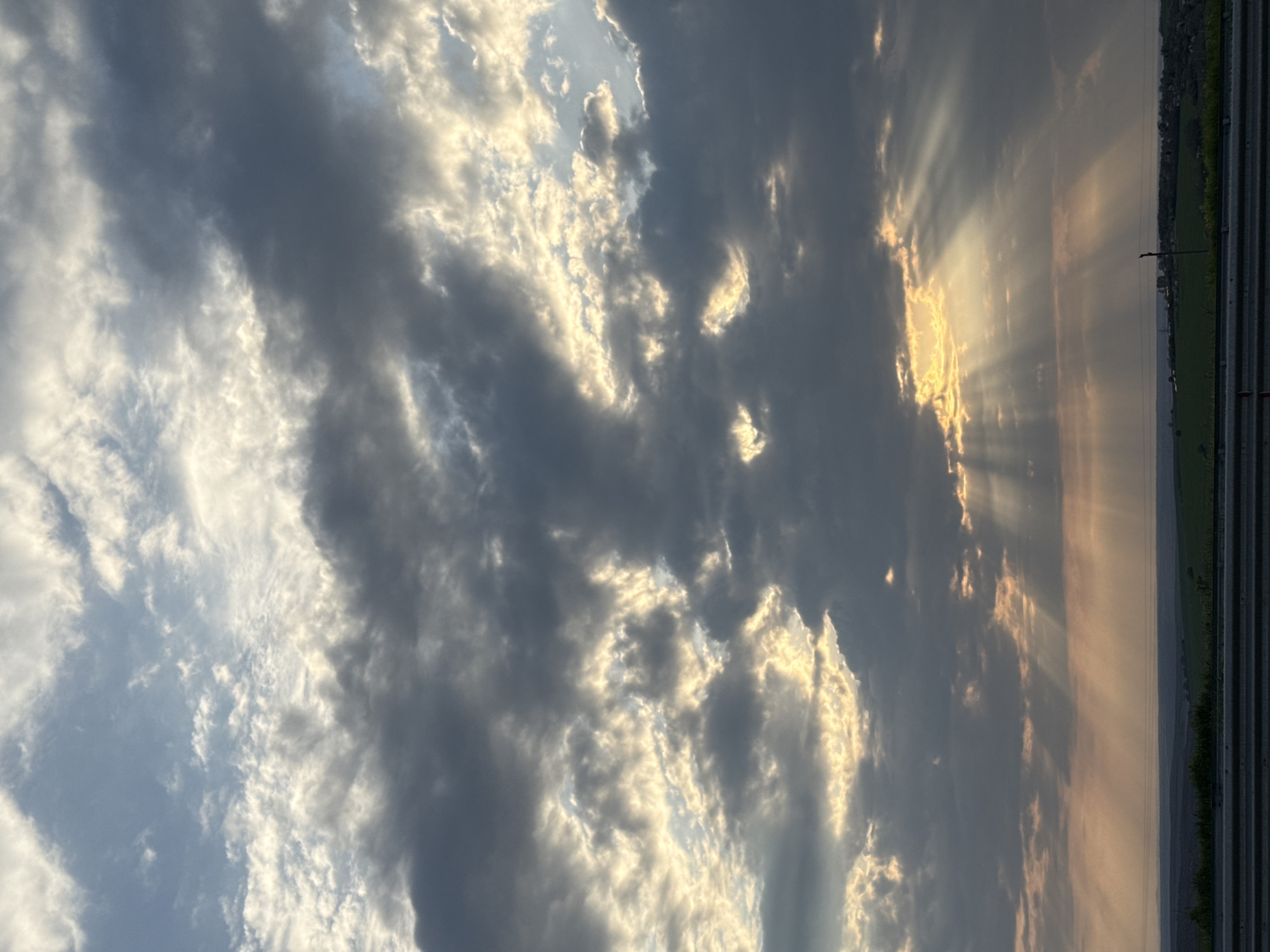 The image shows a dramatic sky with a mix of dark and light clouds. Sunlight breaks through the clouds in beams, creating a dynamic contrast of light and shadow. On the right side of the image, the horizon is visible with a glimpse of land, possibly with some buildings or structures. The location is Pleven, Bulgaria, although specific landmarks are not discernable in this view, focusing primarily on the sky and its conditions.