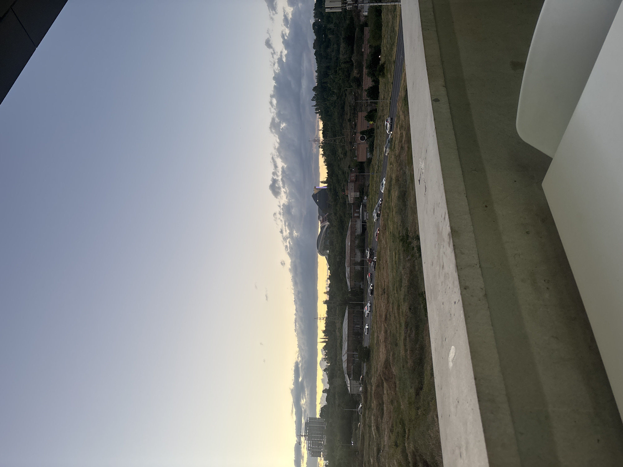 The image captures a panoramic view of an urban landscape, likely at sunset or sunrise, given the warm golden light on the horizon. The sky transitions from a light blue at the top to a pale yellow closer to the horizon, where a distinct band of dark, elongated clouds stretches across the sky.
In the middle ground, the terrain consists of rolling hills covered densely with green trees and some drier, grassy patches. Scattered among the greenery are various buildings, including what appears to be residential structures, some industrial-looking complexes, and a multi-story building under construction, identifiable by a visible crane. A power line pylon stands prominently against the sky near the center of the horizon.
A multi-lane road runs through the landscape, populated with several cars, some of which appear to be parked or moving slowly. The road parallels the vegetated hillsides.
In the foreground, a prominent concrete barrier or ledge occupies the right side of the frame, suggesting the photo was taken from an elevated position, such as a bridge, balcony, or upper floor. Part of a smooth, white, curved architectural element is also visible in the extreme right foreground, partially obscuring the view below.
No people are visibly identifiable in the image, and no legible text can be discerned on any buildings or signs. The scene conveys a tranquil, expansive view of a city's periphery during the golden hour.
