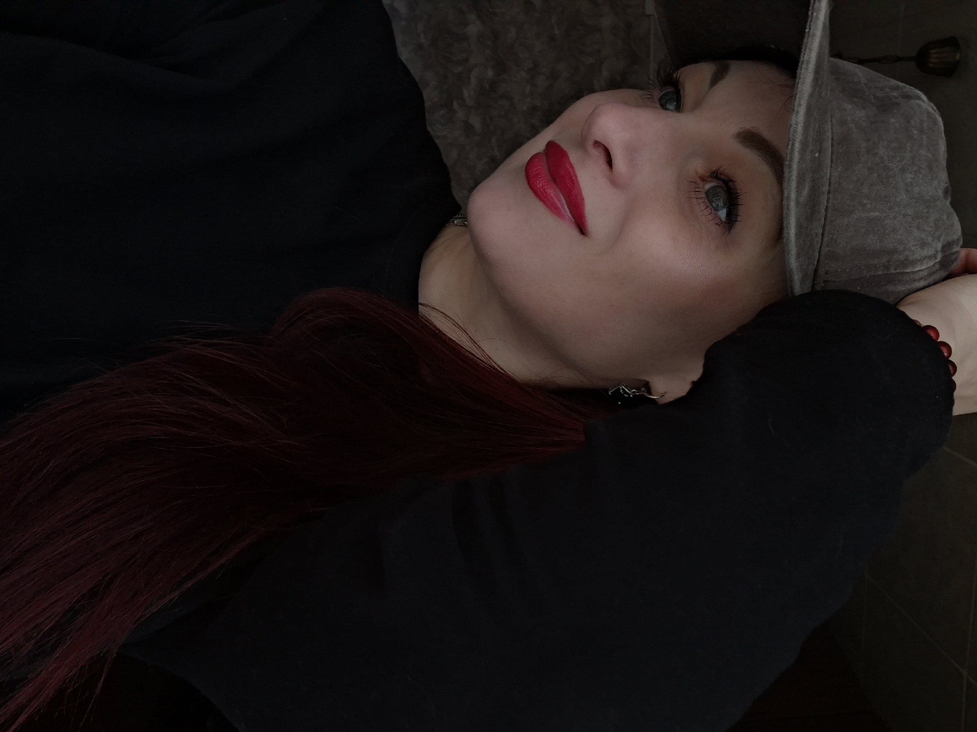 A woman with red hair and blue eyes, wearing a gray hat, is lying down and looking up. She has red lipstick on. The location appears to be indoors. The context of the scene is not clear. The location is in Забалоцкі сельскі Савет, Belarus.