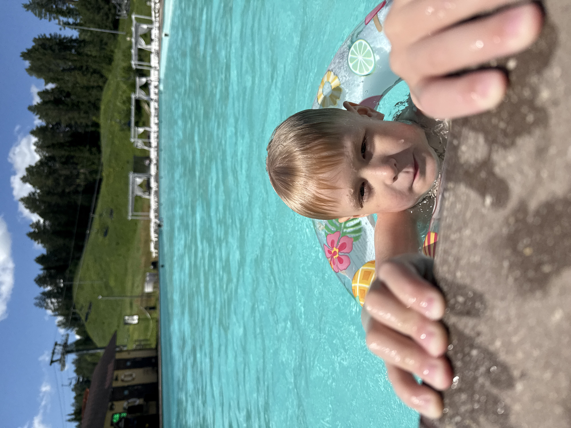 In Nechkino, Russia, a young boy with blonde hair and a floatie is in a bright turquoise swimming pool. He is leaning on the edge of the pool, gripping it with his hands. The floatie has colorful tropical fruit and flower designs. In the background, a grassy hill with pine trees is visible. Ski lifts and a building with a dark roof are also present on the hill. Several white cabanas with draped fabric are lined up along the pool area. The sky is blue with scattered white clouds.