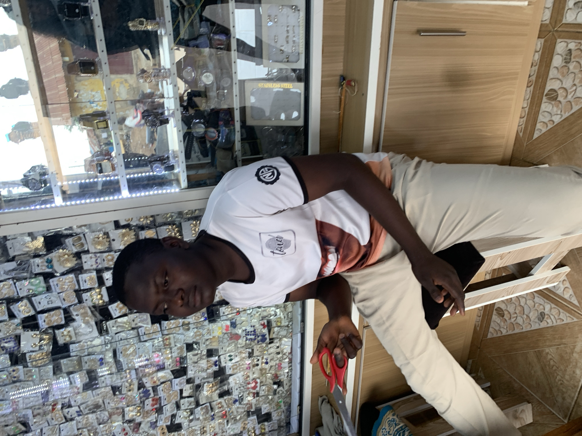 In Abuja, Nigeria, a young Black male sits on a stool in a retail establishment. He is wearing a white t-shirt with a graphic print of lips and a black trim around the collar and sleeves. He is also wearing light-colored pants. In his right hand, he holds a pair of red-handled scissors. The background of the image displays shelves stocked with various items. To the left, shelves are filled with numerous pairs of earrings. To the right, a display case contains watches and other accessories, with an outdoor scene visible through the glass. The flooring is tiled.