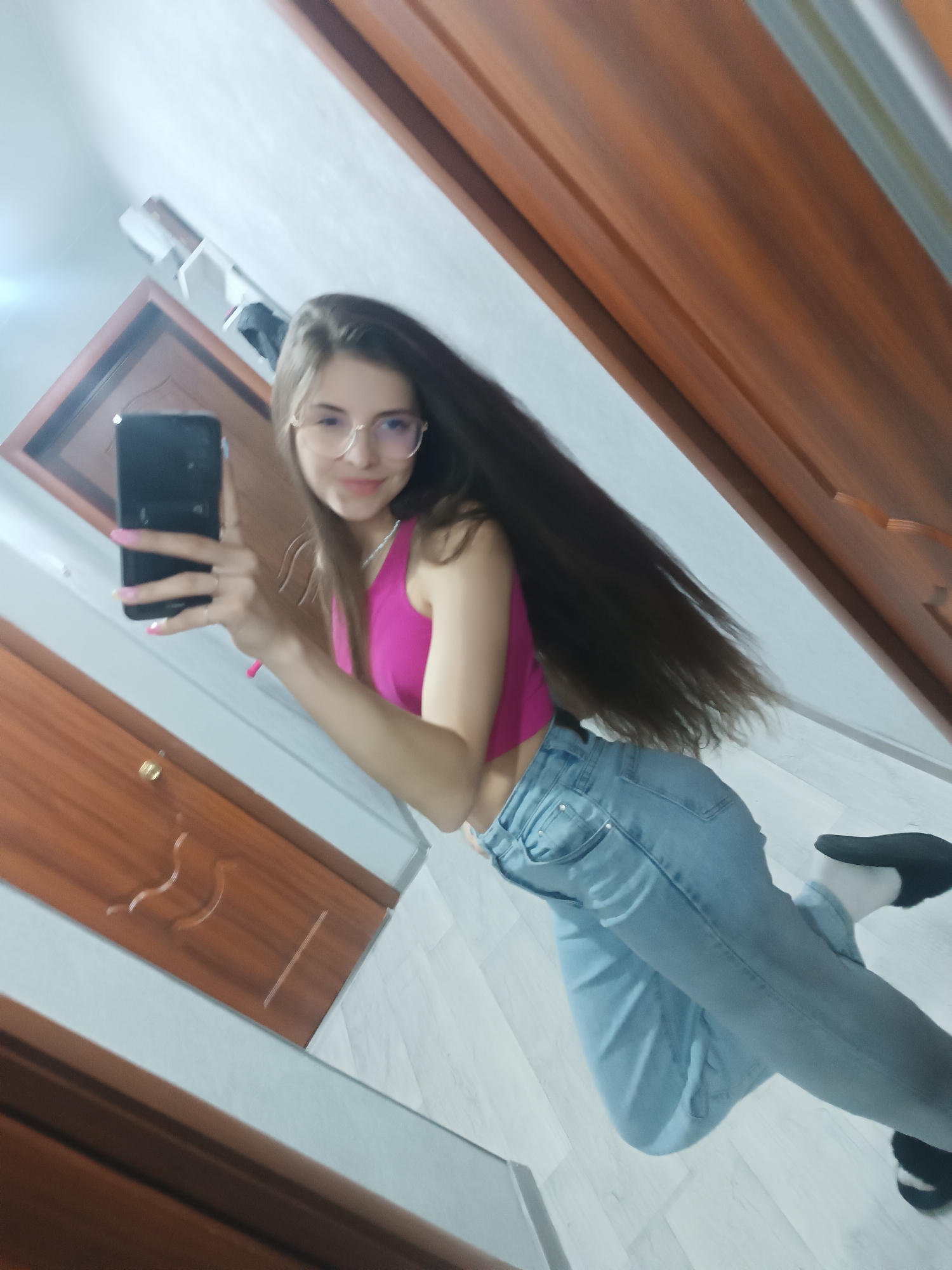 A woman takes a selfie in a room. She is fair-skinned with long brown hair and wearing wire-rimmed glasses, a pink top, and light blue jeans. She holds a black smartphone with pink fingernails visible. She stands in front of a door, the floor is white, and the room's lighting is bright. The location is Chaikovsky, Russia.