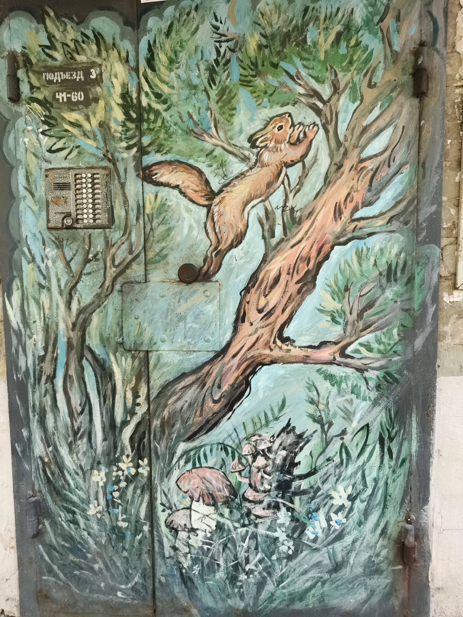 A painted metal door depicts a forest scene with a squirrel climbing a tree. The squirrel is depicted in mid-leap, reaching towards a branch. At the base of the tree, there are mushrooms and small white flowers. The background is a mix of green foliage and light blue sky. On the left side of the door, there is an intercom panel and a sign with Cyrillic text that reads "ПОДЪЕЗД 3" and "41-60", indicating entrance 3, apartments 41-60. This entrance is located in the city of Chaikovsky, Russia. The door itself is weathered and shows signs of rust and paint wear. The overall style of the painting is naive and colorful.