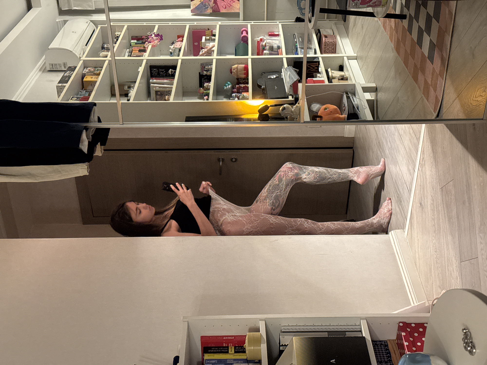 The image shows a young woman standing indoors, leaning against a wall and looking at her phone. She is wearing a black tank top and white lace tights. Her left leg is bent, and her right leg is extended. She has tattoos on her right leg. To her right, a tall, white bookshelf is visible, filled with various items including books, boxes, and decorative objects. A large mirror reflects part of the room, showing the bookshelf and a rug on the floor. The room appears to be a living space or bedroom, with light-colored walls and wooden flooring. The overall setting suggests an interior environment. The image does not contain any explicit information about the location being Moscow, Russia.