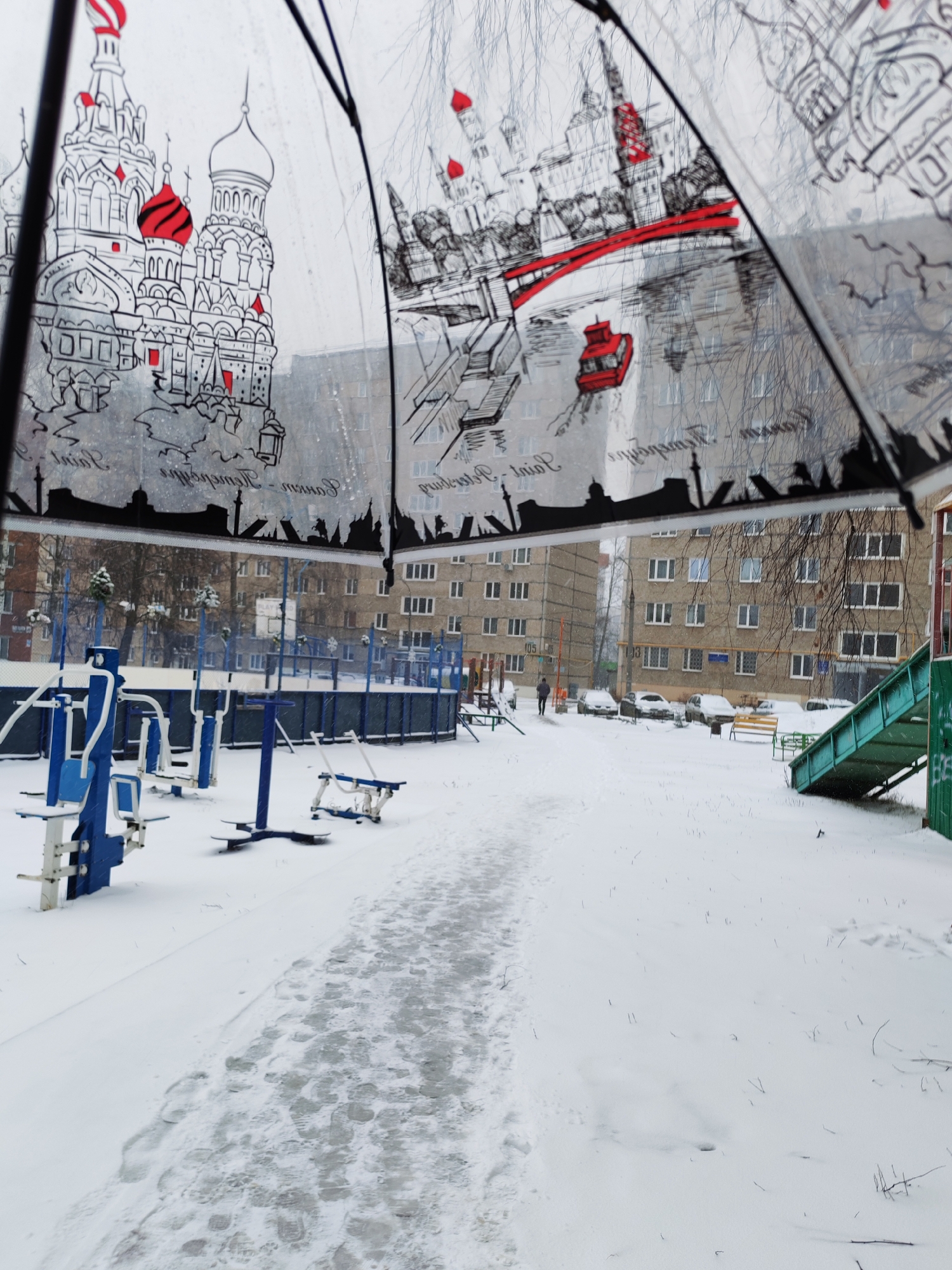 The image captures a snowy scene in Izhevsk, Russia, as viewed from under an umbrella. The umbrella is transparent with a black-and-white drawing of architectural landmarks and a river scene with red details. The scene outside the umbrella depicts a winter day with buildings and a playground covered in snow. The ground is mostly white, and some footprints are visible on a partially cleared path. Exercise equipment, a slide, and a fenced-in sports area are visible in the foreground. In the background, apartment buildings line the street, and cars are parked on the side. A lone figure is walking in the distance.