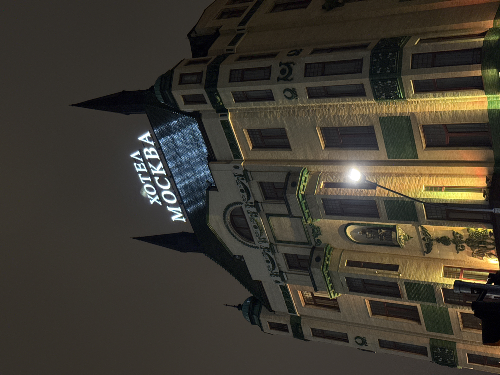 A low-angle, nighttime shot shows the Hotel Moskva in Belgrade, Serbia. The building's facade is illuminated with warm light, highlighting its architectural details. The hotel's name, "HOTEL MOSKVA," is displayed in bright, vertical white lettering on one of the building's angled surfaces. The sky is dark. A streetlight is visible in front of the building. A portion of a vehicle is visible in the lower right corner.