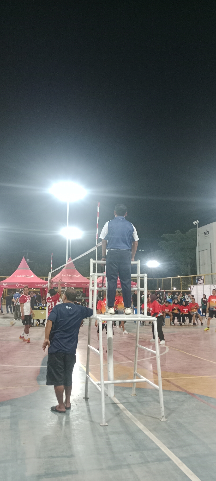 In an outdoor volleyball court at night, illuminated by bright floodlights, a volleyball match is in progress. The court surface is marked with colored lines. A referee stands on a tall white platform, observing the game. On the court, players in red and white uniforms are actively playing. Spectators are seated or standing around the perimeter of the court, many wearing red shirts. Red tents are set up behind the court, bearing the "bankjatim" logo. A building with the "SIG" logo is visible in the background. This scene is located at the Tuban Sport Center, Tuban, East Java, Indonesia.