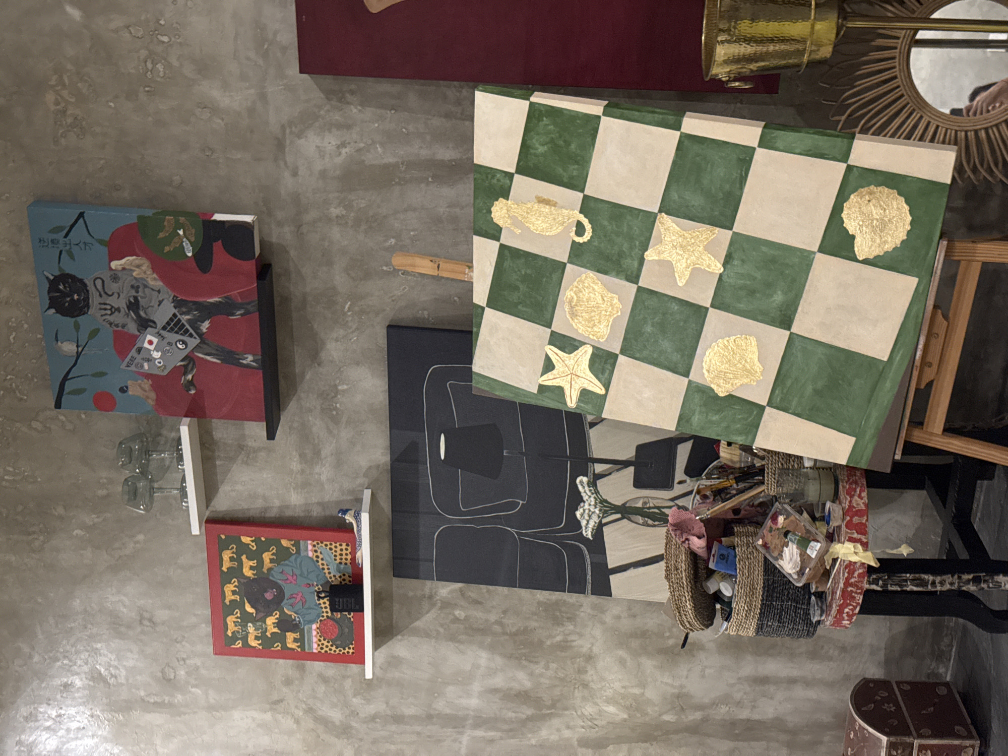 The image depicts an indoor setting in North Kuta, Indonesia, showcasing a collection of art and decorative objects arranged on a wall and table. The wall displays several framed paintings, including one with a cat-like figure and text in Japanese, and another with elephant motifs. Below the paintings, glass objects are visible. A chessboard with alternating green and beige squares is propped up on an easel. Various golden shapes, including a genie lamp, starfish, and seashells, are affixed to the chessboard. A golden telescope-like object sits near a decorative mirror.
Below the chessboard is a black painting of a chair next to a basket with art supplies, including brushes, paint tubes, and containers. A table with red and white patterns supports the art supplies. In the background, a wooden trunk with decorative patterns is visible. The setting appears to be an art studio or display space.