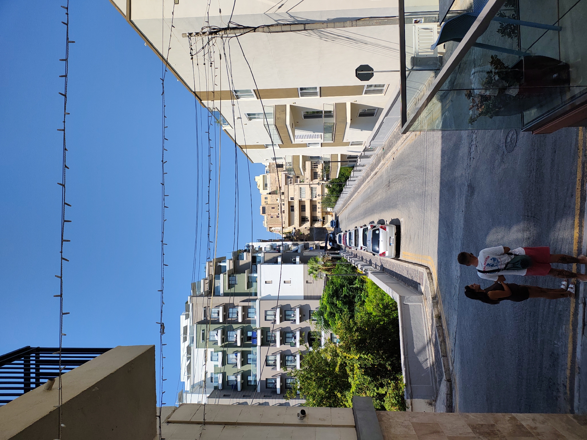 In the city of Manikata, Malta, on a sunny day, a street is lined with modern apartment buildings. The street slopes gently uphill and is populated by parked cars, mostly white. On the right side of the street, a modern white apartment building features balconies with glass railings. On the left, another apartment building is visible, characterized by its green and white facade and numerous balconies. In the foreground, two individuals, a woman and a man, stand on the street, facing away from the camera. The woman wears a dark top and shorts, while the man is in a white t-shirt, red shorts, and a green bag slung across his shoulder. Overhead, decorative string lights are strung across the street. The sky is a clear, vibrant blue.