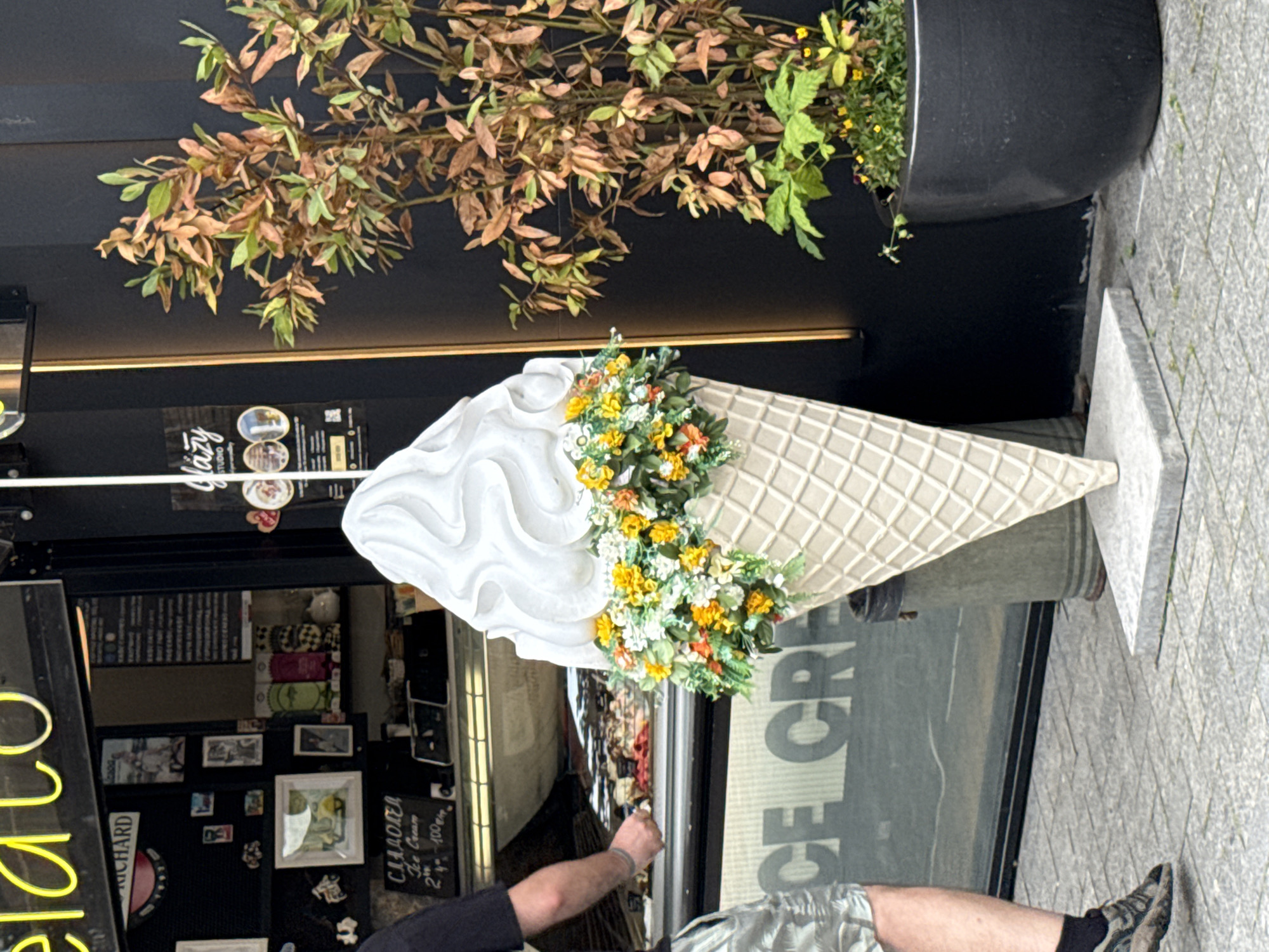 A person wearing shorts and dark socks with athletic shoes is partially visible on the left side of the image. They are standing on a paved sidewalk next to a large, white ice cream cone sculpture decorated with artificial flowers in orange, yellow, and white. Behind the sculpture, a dark wall is visible, with a plant in a large black planter to the right. In the background, the exterior of an establishment with a window displaying ice cream tubs is visible. A neon "gelato" sign is partially visible at the top of the image. The word "ICE CREAM" is stenciled in large letters on the glass window.
