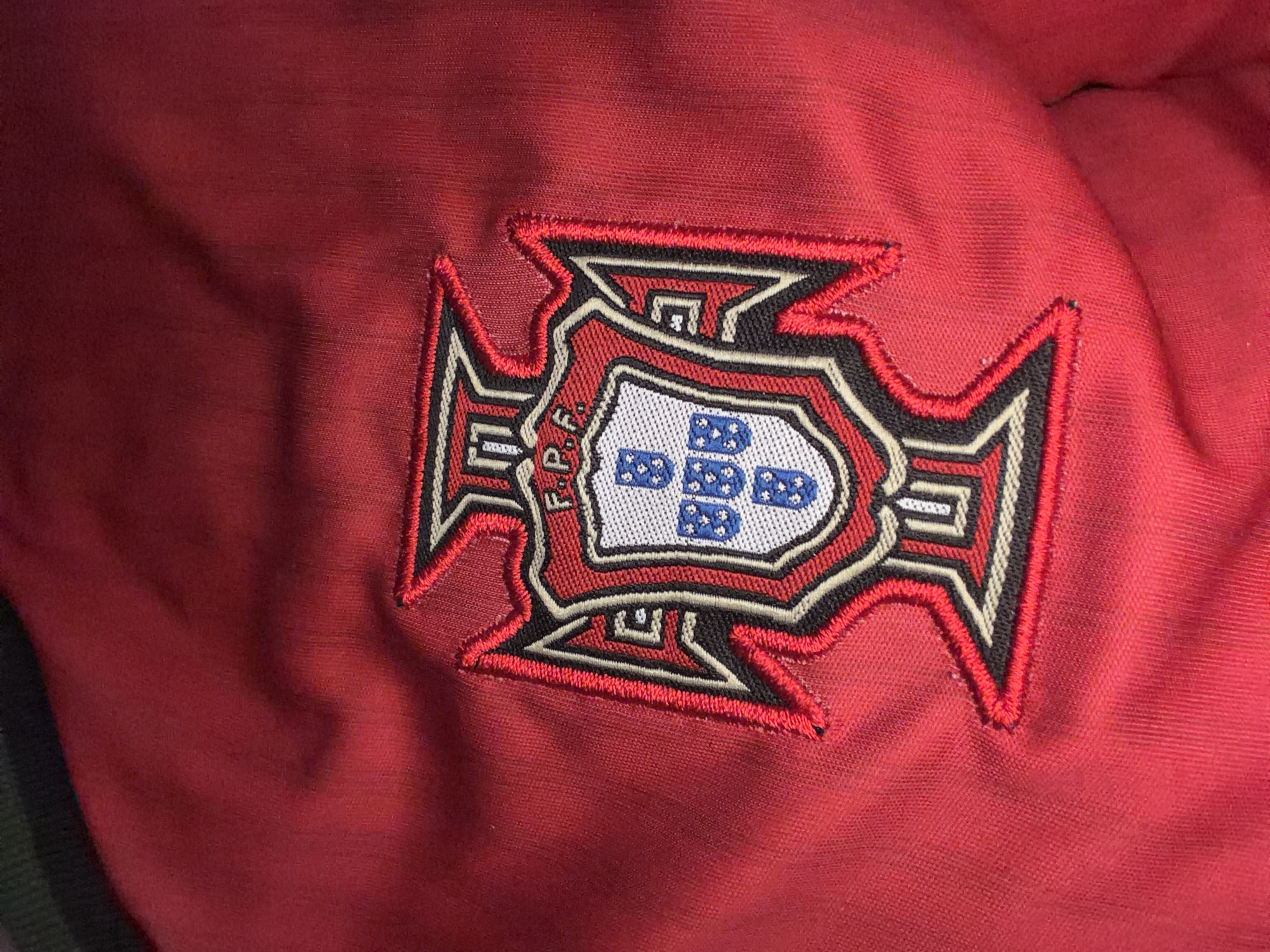 The image shows a close-up of an embroidered emblem on a red fabric. The emblem is shield-shaped with ornate borders and features a smaller shield in the center. The central shield contains five blue escutcheons, each with five white bezants. Above the central shield, the letters "F.P.F." are visible in white embroidery. The outer shield is outlined in red, black, and white embroidery, with stylized angular shapes extending outwards. The red fabric appears to be a garment, possibly a jacket or shirt, with visible stitching and texture. The lighting in the image creates some shadows and highlights on the fabric, emphasizing its texture and the dimensionality of the embroidery. The background is out of focus, showing more of the red fabric. No specific location in Nikopol, Bulgaria, can be determined from this image alone.