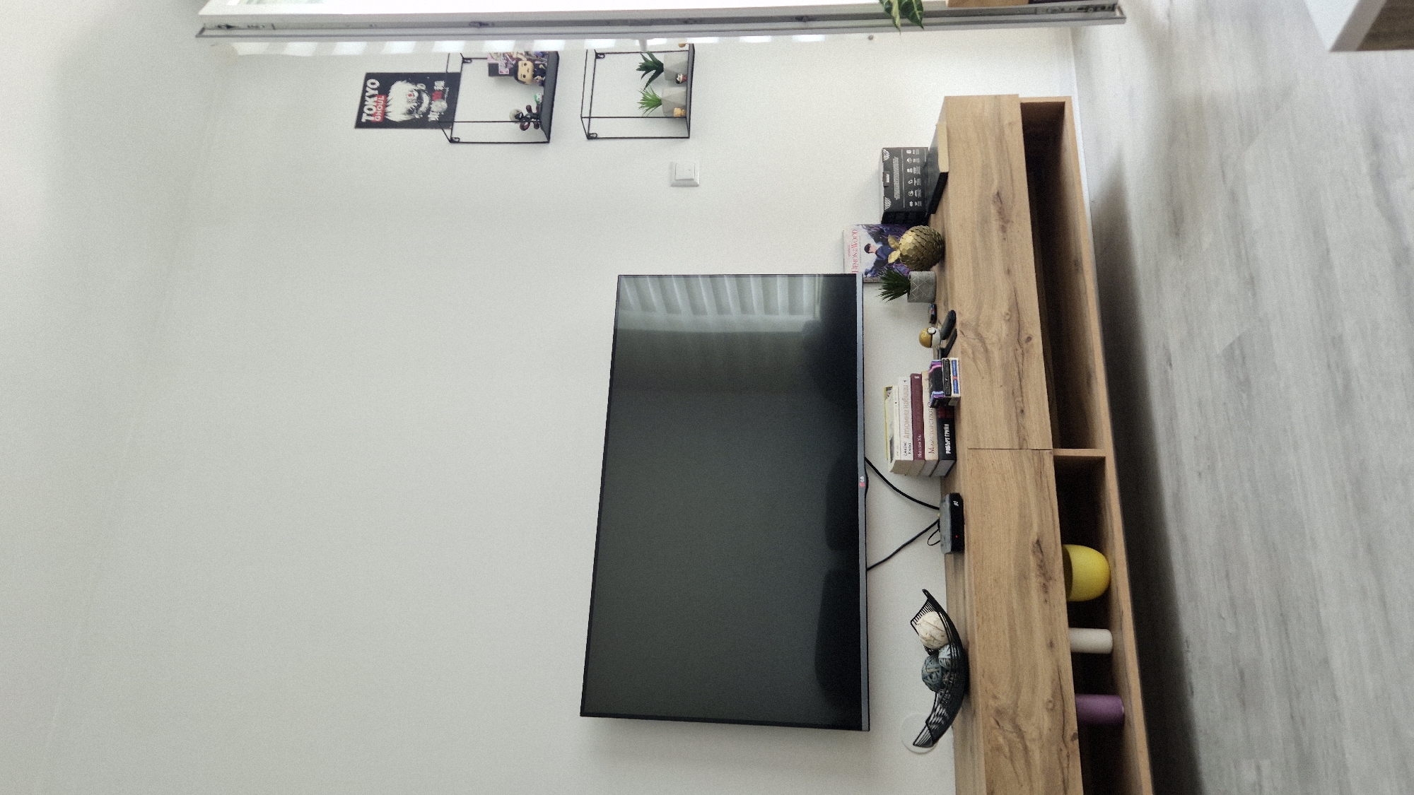 The image shows an indoor scene. A black flat-screen television is mounted on a white wall, left of a light wood grain cabinet. Shelves above the TV are decorated with a Tokyo Ghoul poster, a figurine in a black frame, and greenery in another frame. A light switch is between the shelves. On top of the cabinet are assorted decorative objects, books, and electronics. The floor is light gray and the ceiling is visible at the top. The image does not specify Burgas, Bulgaria.