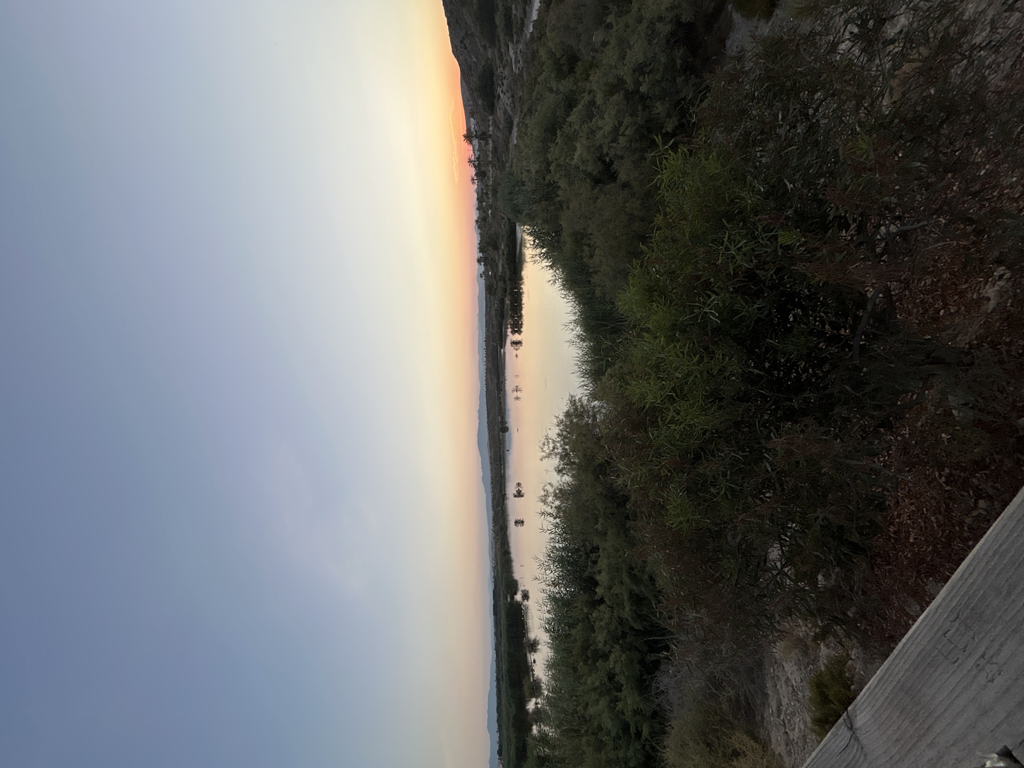 The image depicts a tranquil natural landscape at dusk. A still body of water, likely a lagoon or salt flat, dominates the mid-ground, reflecting the soft hues of the fading sky. Surrounding the water is dense, green vegetation, appearing as bushes and reeds. In the background, a range of low hills is visible, silhouetted against the horizon. The sky itself transitions from a clear, pale blue at the top to a warm gradient of yellow and orange near the horizon, indicating the setting sun. In the foreground, a wooden railing is partially visible on the left, suggesting a viewpoint or walkway. The overall scene is serene and untouched, characteristic of a natural preserve or undeveloped coastal area. The location is identified as the Laguna de Puertollano in Vera, Spain, a protected wetland area known for its diverse birdlife and unique ecological features. There are no people or discernible human activity in the image, emphasizing the natural state of the environment.