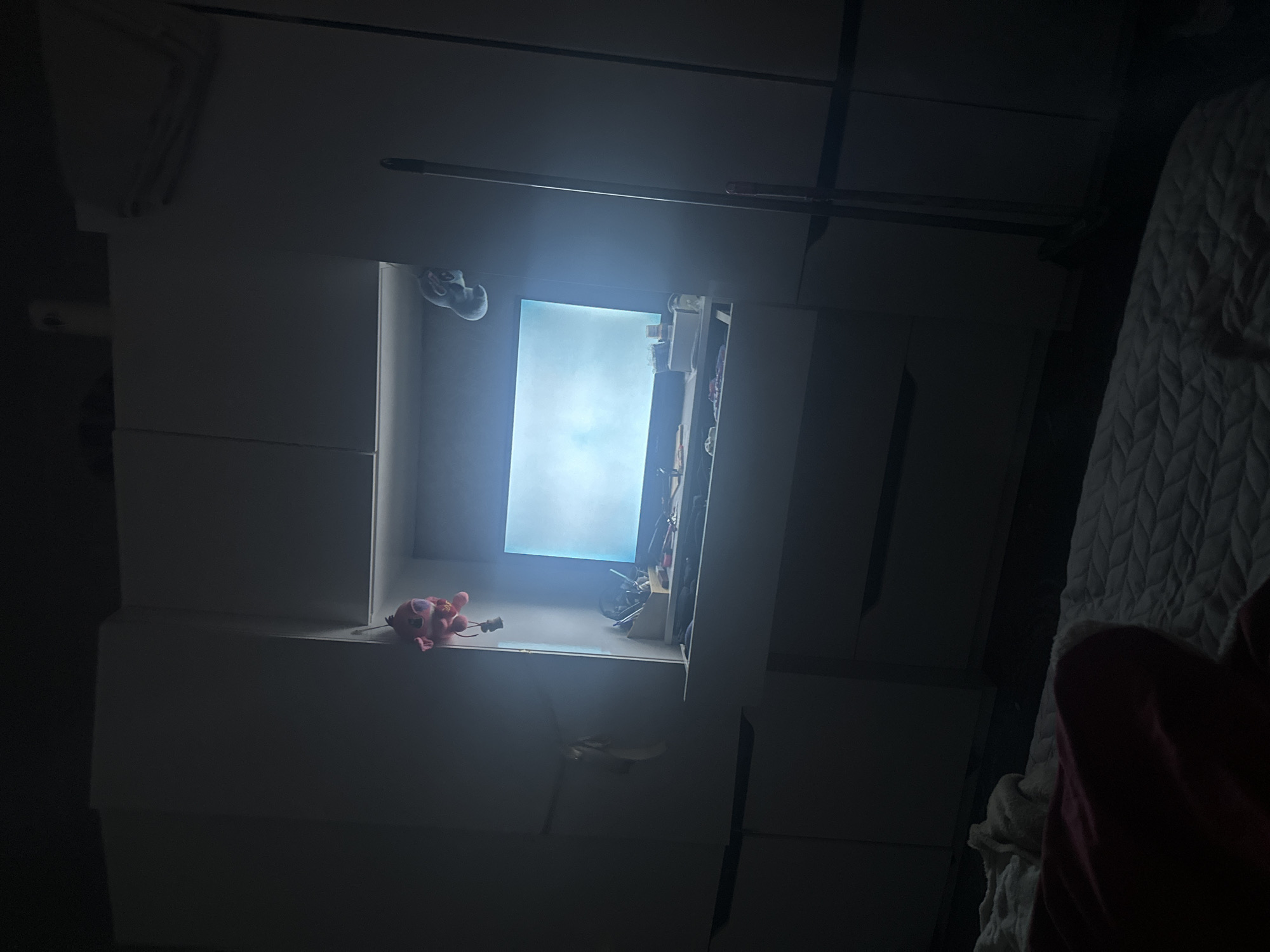 The image depicts a dark, interior view, possibly a bedroom, with a focus on a white shelving unit. The shelving unit has open compartments containing various items, including a pink stuffed animal, shoes, and other miscellaneous objects. A bright light source, likely daylight, is visible through a window or opening in the shelving unit. A bed is partially visible on the right side of the frame, covered with a textured white and gray blanket and a red garment. The overall environment appears cluttered and dimly lit. There is no definitive information to place the location within the city of Sofia, Bulgaria.