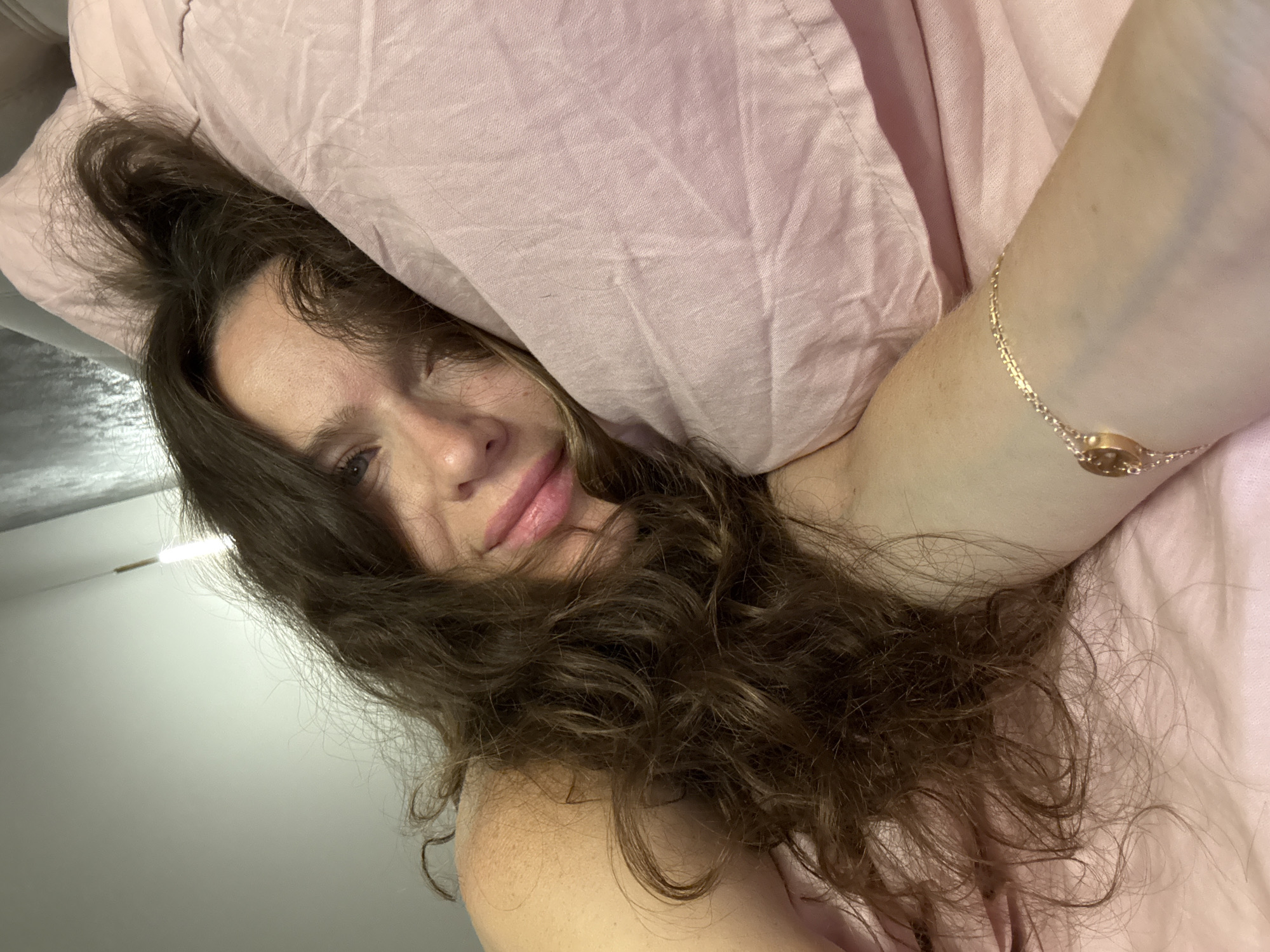 A woman with long, wavy brown hair is lying in bed, resting her head on a pink pillow. Her eyes are open and she is looking at the camera. She is wearing a pink bracelet on her left wrist. The background is a bed with pink sheets and a pillow. The image appears to be taken indoors in a bedroom. There is no specific information available about the location in the city of Dolgoprudny, Russia.