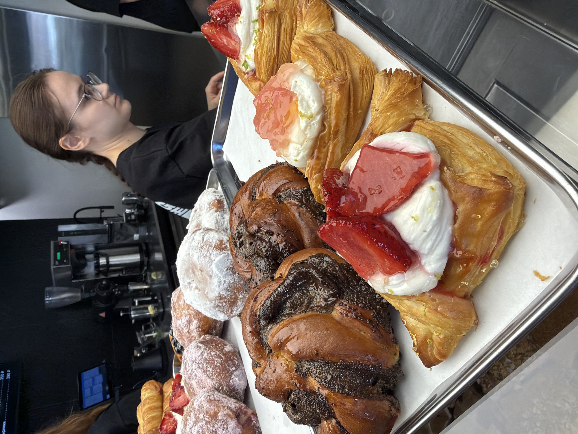 The image shows a display case filled with pastries in what appears to be a bakery or cafe. In the foreground, there are several flaky pastries topped with cream and fruit. One pastry has sliced strawberries and a red glaze, while another is topped with a dollop of white cream and what looks like a pink jelly or fruit compote with flecks of green garnish. Behind these, there are darker, swirled pastries with a glossy finish, possibly filled with chocolate or poppy seeds. Further back, more pastries, some covered in powdered sugar, are visible.
A young woman with her hair in a braid and wearing glasses and a black t-shirt is standing behind the counter, partially visible on the right side of the image. She is looking down towards the pastries, seemingly attending to them. The background shows stainless steel appliances, likely a coffee machine and other cafe equipment, against a dark wall. The overall impression is one of a well-stocked pastry display. The context provided indicates this scene is located in Izhevsk, Russia.
