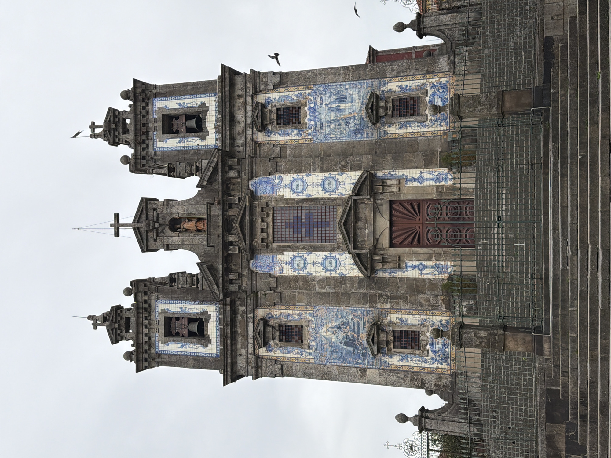 This image depicts the Igreja do Carmo, a notable church in Porto, Portugal. The church's facade is adorned with extensive azulejo tiles, a traditional Portuguese ceramic tile art form. The tiles primarily feature a blue and white motif, depicting various scenes and ornamental patterns. The architecture is a blend of Baroque and Rococo styles, characterized by its grand scale, ornate stonework, and prominent bell towers.
The scene is captured from a slightly elevated perspective, looking up towards the church entrance and its flanking towers. A large set of stone stairs leads up to the church. The sky is overcast, suggesting a cloudy or perhaps misty day, common in Porto. There are no people visible in the frame, giving the image a sense of quiet grandeur. The overall mood is one of solemnity and historical reverence, highlighting the architectural beauty and cultural significance of the church.
Notable details include the intricate stonework around the windows and doorways, the decorative finials topping the towers, and the wrought-iron fencing that partially encloses the approach to the church. Faint text is visible on some of the azulejo panels, though it is too small and blurry to be definitively read. The image's orientation is vertical, with the top of the church appearing to the left.