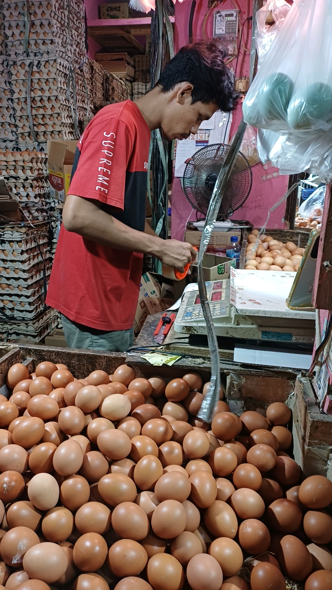 In Citeureup, Indonesia, a man in a red and black t-shirt stands in a market stall filled with numerous brown eggs. He is holding an orange object, possibly a tool, and appears to be interacting with a scale. Stacks of egg cartons are visible in the background, along with a fan and other market goods. The scene captures a moment of commerce within a bustling local market.