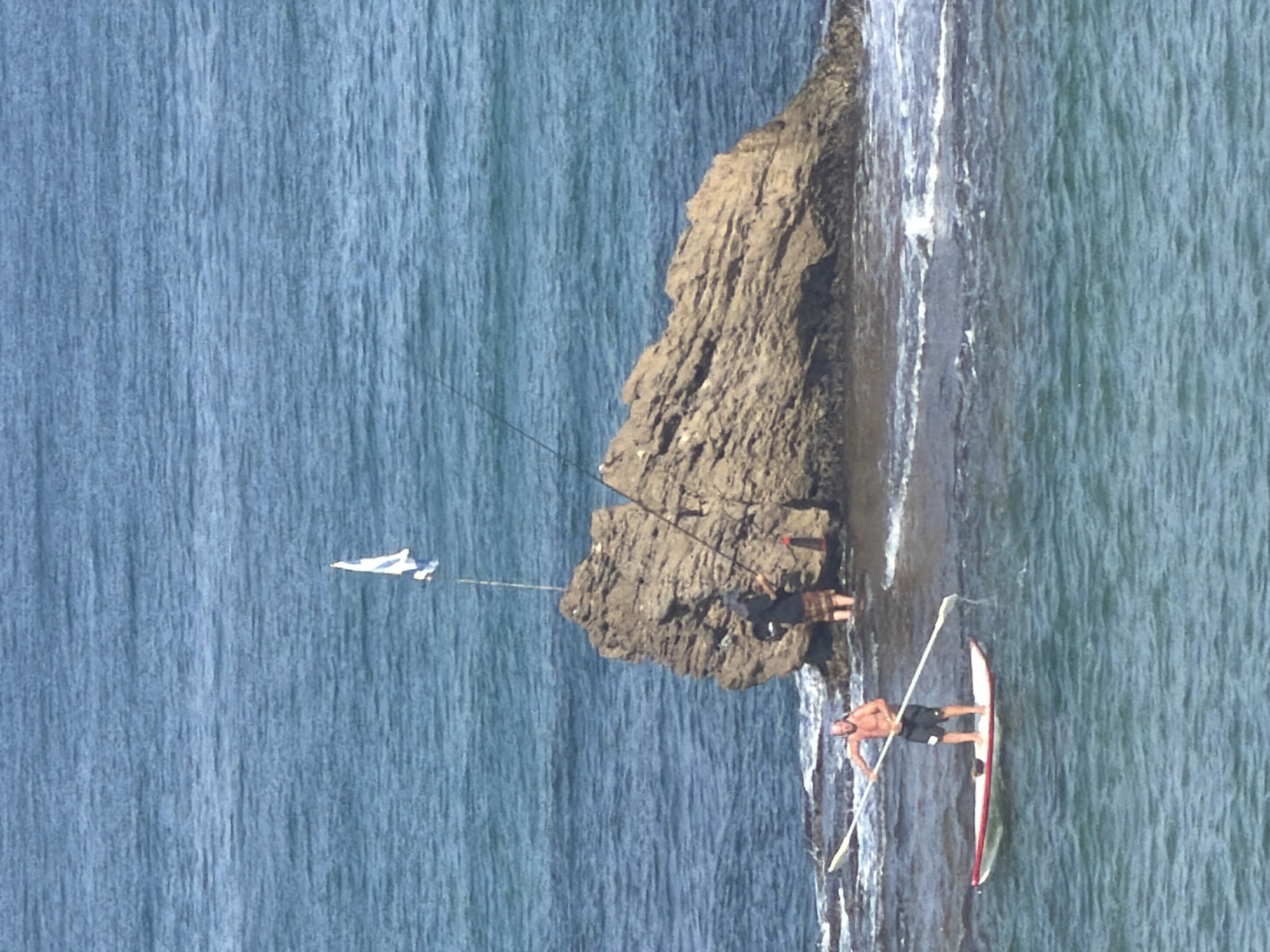 The image depicts a rocky coastline in Bat Yam, Israel, under a clear, bright sky. The ocean, a deep blue, stretches across the frame, with gentle waves lapping against the shore and around submerged rocks.
In the foreground, a man stands on a red and white paddleboard, holding a long paddle horizontally. He is shirtless and wearing dark shorts. The paddleboard is positioned in relatively calm, shallow water.
Further back, a large, weathered rock formation rises from the sea. On top of this rock, a person wearing a dark shirt and a plaid skirt-like garment is visible, appearing to be fishing. A fishing rod extends from their hand into the water. A small white flag with blue markings is mounted on a pole atop the rock formation. Another fishing rod is also visible, extending from the rock towards the right side of the image.
The water around the rock is churned with white foam from incoming waves. The overall scene suggests a recreational day at the beach, with individuals engaged in water sports and fishing activities.
